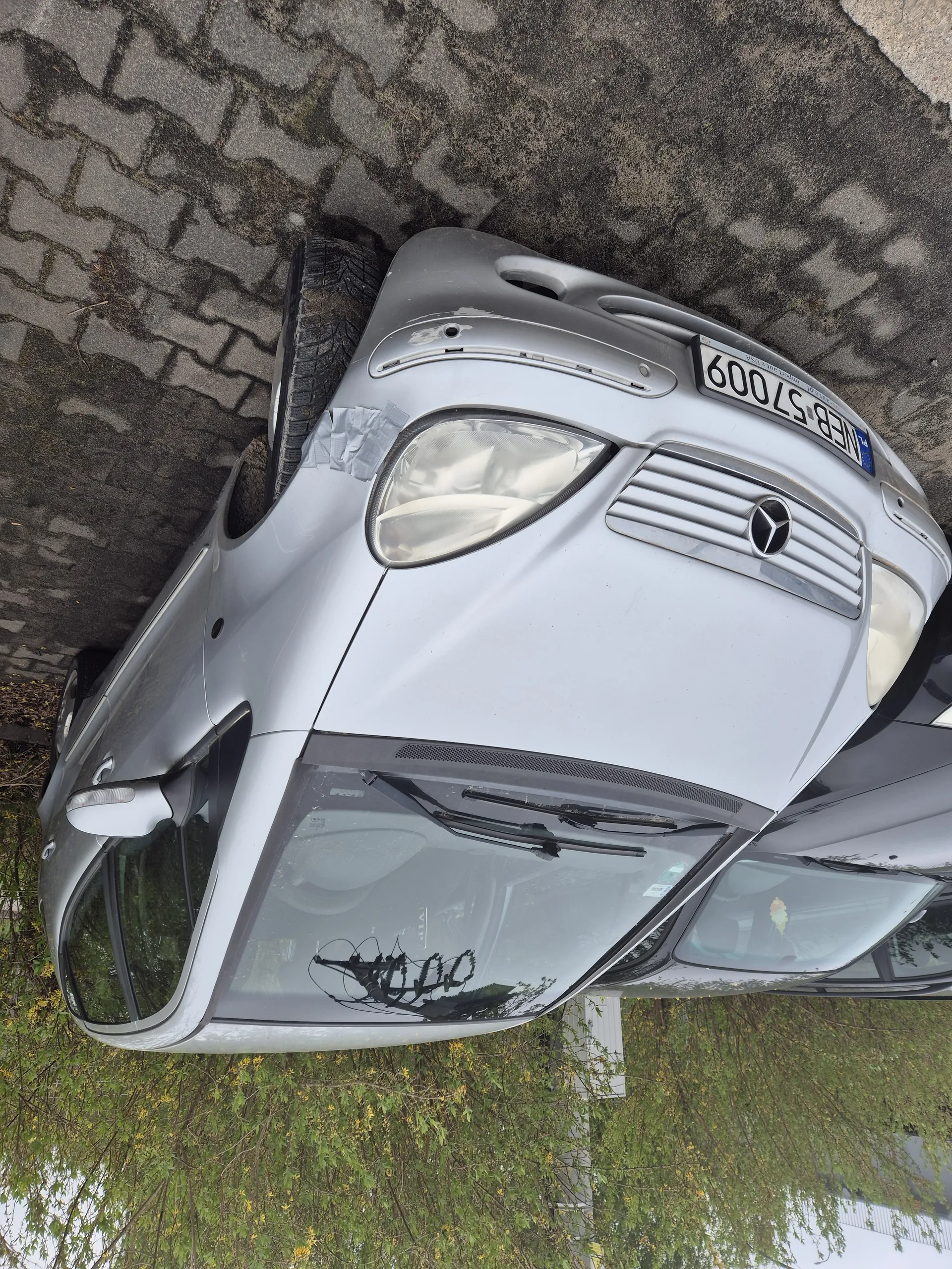Samochód marki Mercedes-Benz w kolorze srebrnym zaparkowany na brukowanej powierzchni. Widoczna tablica rejestracyjna WEB 57009. Na przedniej prawej stronie pojazdu uszkodzenia. W tle krzewy z żółtymi kwiatami oraz fragment innego czarnego samochodu.