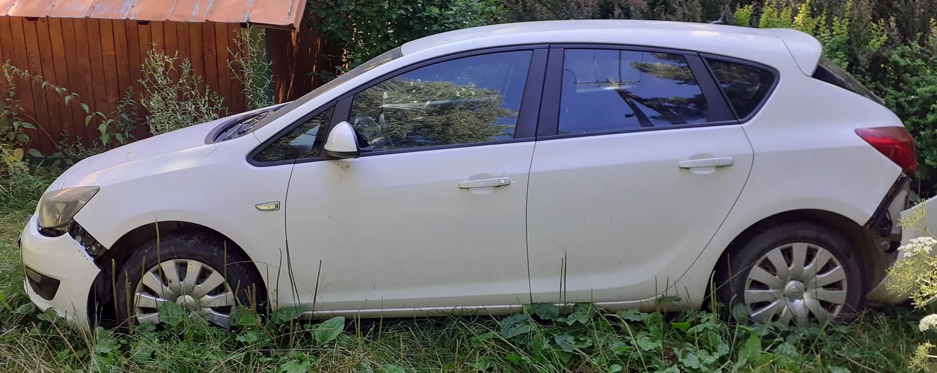 Boczny widok białego samochodu osobowego typu hatchback, zaparkowanego w wysokiej trawie na tle drewnianego budynku i zieleni. Pojazd posiada widoczne uszkodzenia w przedniej części, w okolicach lewego reflektora i zderzaka.