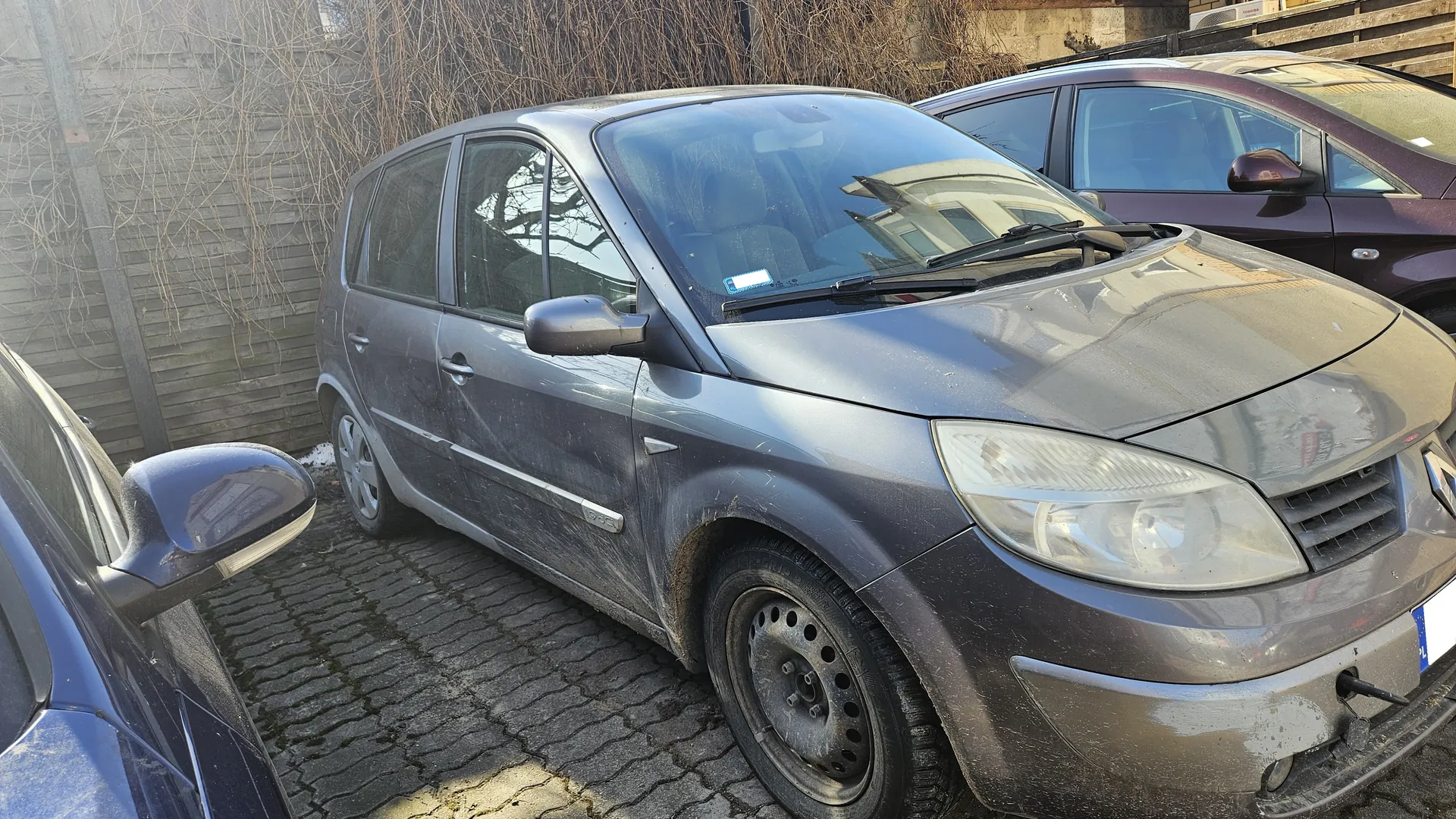 Szary samochód osobowy zaparkowany na brukowanej powierzchni. Pojazd jest zabrudzony, z widoczną warstwą kurzu i błota na karoserii oraz felgach. W tle widoczne są inne samochody: niebieski po lewej stronie i bordowy po prawej. Za pojazdami znajduje się drewniane ogrodzenie oraz suche pnącza. Na przedniej szybie samochodu umieszczony jest biały znaczek. Tablica rejestracyjna na przodzie jest częściowo widoczna, ale tekst nieczytelny. Oświetlenie wskazuje na słoneczny dzień.