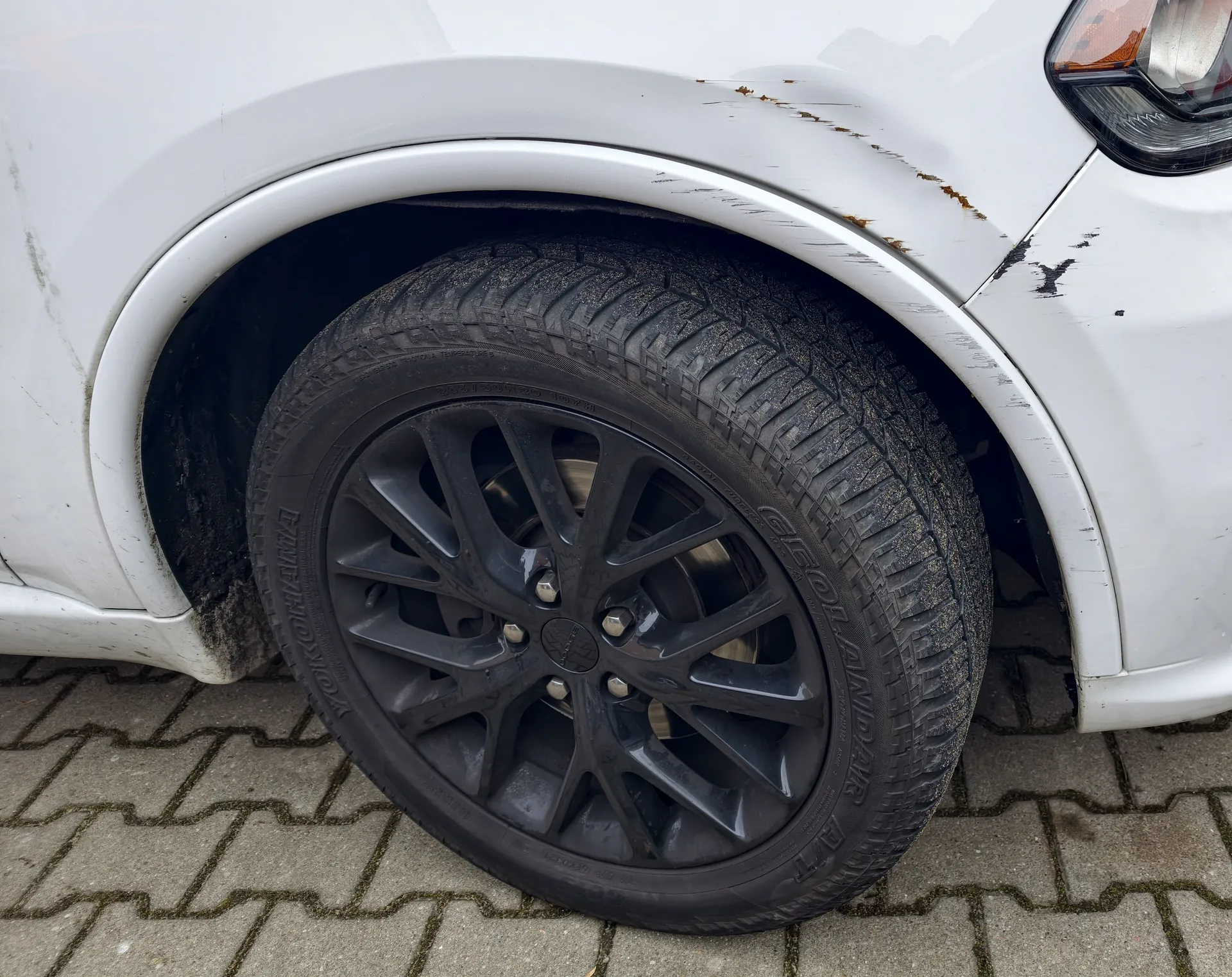 Biały samochód z widocznym przodem koła. Czarna felga aluminiowa z oponą zimową o wyraźnym, głębokim bieżniku. Na karoserii w okolicy nadkola widoczne ślady brudu i zadrapań. Samochód stoi na brukowanej powierzchni z kamiennymi płytami ułożonymi w geometryczny wzór. Część przodu pojazdu, w tym reflektor, jest widoczna po prawej stronie obrazu.