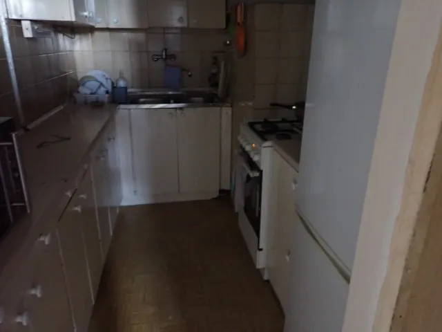 Wnętrze małej, jasnej kuchni z białymi szafkami kuchennymi, blatem i sprzętami. Po lewej stronie znajduje się długi blat z szafkami pod spodem. W głębi widoczny jest zlew dwukomorowy z baterią oraz szafki ścienne. Po prawej stronie stoi biała kuchenka gazowa z piekarnikiem oraz fragment białej lodówki na pierwszym planie. Ściany wyłożone są jasnymi kafelkami, a podłoga jest w kolorze beżowym.