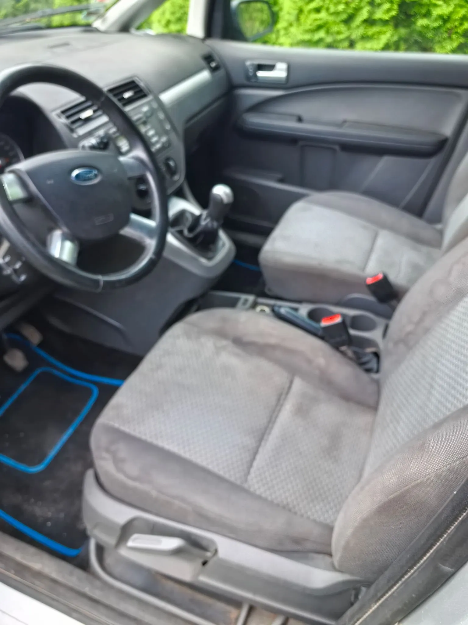 Wnętrze samochodu marki Ford widziane z perspektywy otwartych przednich drzwi pasażera. Widoczna jest kierownica z logo Forda, deska rozdzielcza z radiem i nawiewami, lewarek zmiany biegów oraz dwa szare, materiałowe fotele. Na podłodze znajdują się niebieskie gumowe dywaniki.