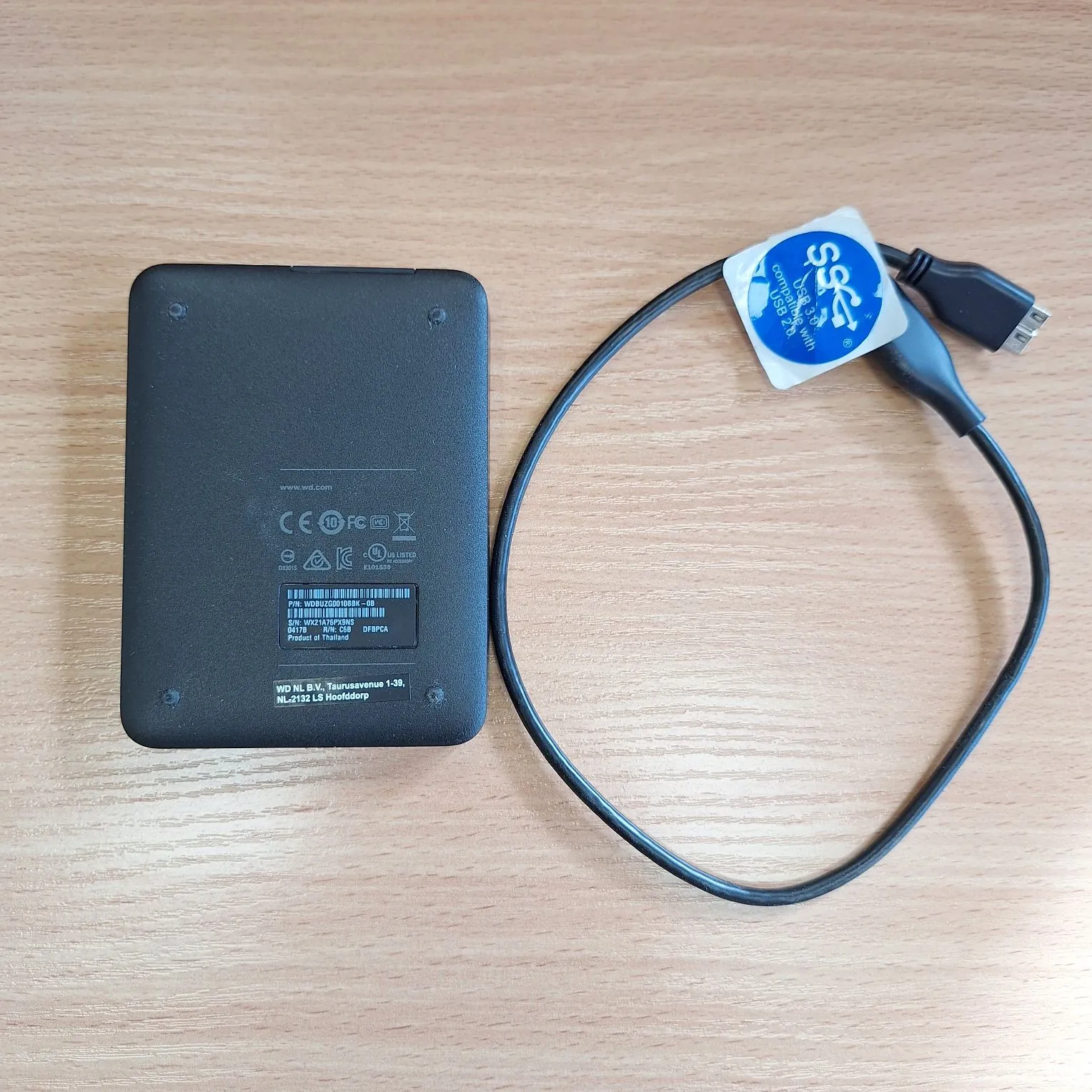Czarny zewnętrzny dysk twardy leżący na drewnianym blacie obok czarnego kabla USB. Na spodzie dysku znajduje się etykieta z informacjami technicznymi, w tym logo CE, FC oraz napisy: 'www.wd.com', 'P/N: WDBWUZ0000BBK', 'S/N: WX1N1TP0PNS'. Kabel posiada niebieską zawieszkę z napisem 'Compatible with USB 2.0'.