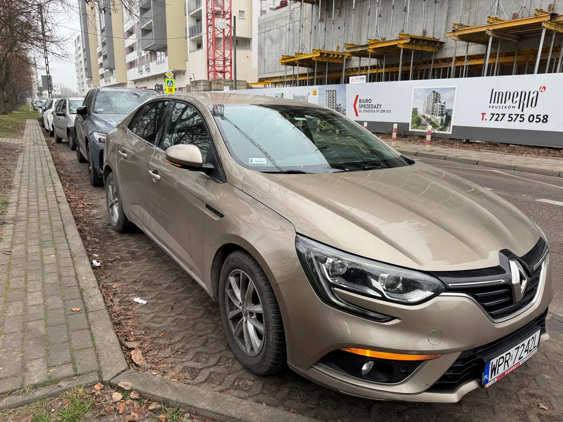 Zdjęcie przedstawia brązowy samochód marki Renault zaparkowany przy chodniku z kostki brukowej. Na pierwszym planie widoczna tablica rejestracyjna WPR 7242L. Za samochodem kolejne zaparkowane pojazdy. W tle budynki mieszkalne, czerwona wieża kranowa oraz budowa z żółtymi rusztowaniami. Na białym banerze widnieje napis Imperia Pruszków, T. 727 575 058 oraz Biuro Sprzedaży ul. Staszica 1F lok. U12. Chodnik pokryty suchymi liśćmi, po lewej stronie drzewa bez liści.