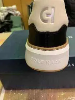 Biało-czarna buty marki Cole Haan widziane od tyłu. Na pięcie widoczny napis 'CH', a na białej podeszwie napis 'COLE HAAN'. W tle niebieskie pudełko i rozmyte przedmioty.