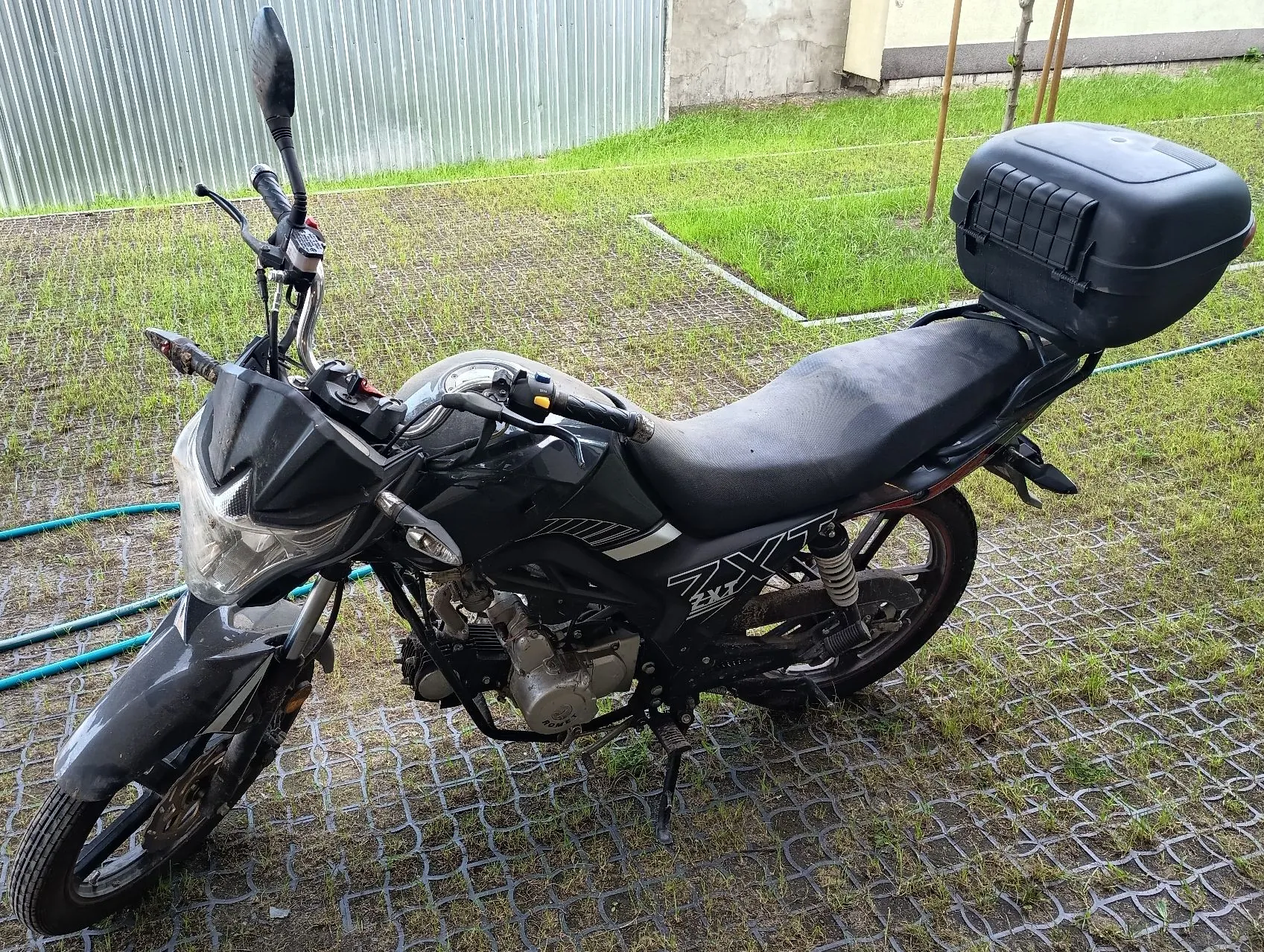 Czarny motocykl zaparkowany na trawniku z betonowymi kratkami. Motocykl posiada czarny kufer zamykany na klucz zamontowany z tyłu oraz napis 'ZX' na boku owiewki. W tle widoczny jest metalowy płot z blachy trapezowej oraz zielona trawa.