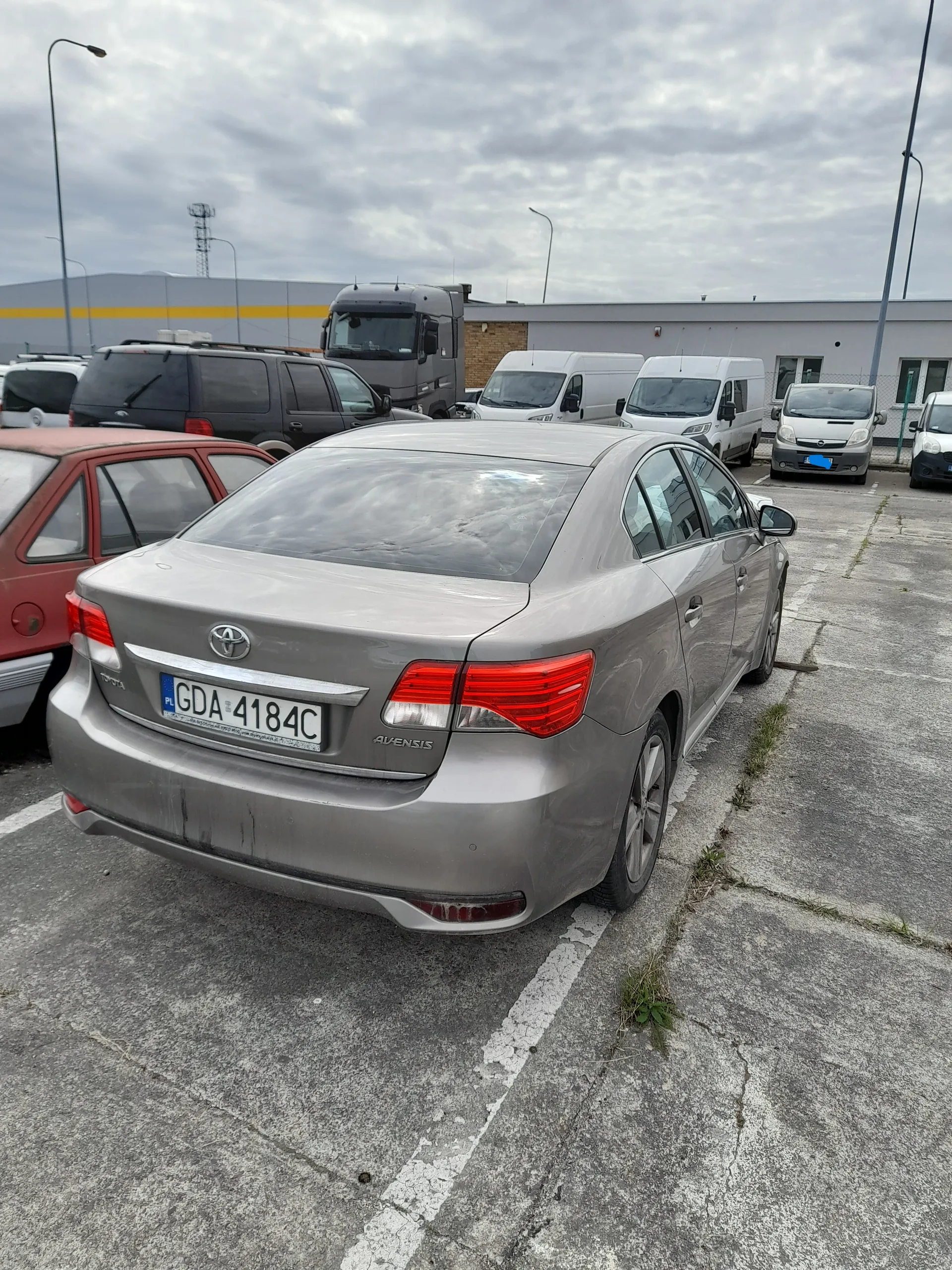 Zdjęcie przedstawia szary samochód osobowy marki Toyota Avensis zaparkowany na betonowym parkingu. Widoczny jest tył pojazdu z tablicą rejestracyjną PL GDA 4184C. W tle znajdują się inne pojazdy: czerwony samochód osobowy, ciemny SUV, białe furgony oraz ciężarówka. Tło stanowi budynek przemysłowy z żółtą listwą, słupy oświetleniowe i zachmurzone niebo. Na parkingu widoczne są białe linie wyznaczające miejsca parkingowe oraz szczeliny z trawą.