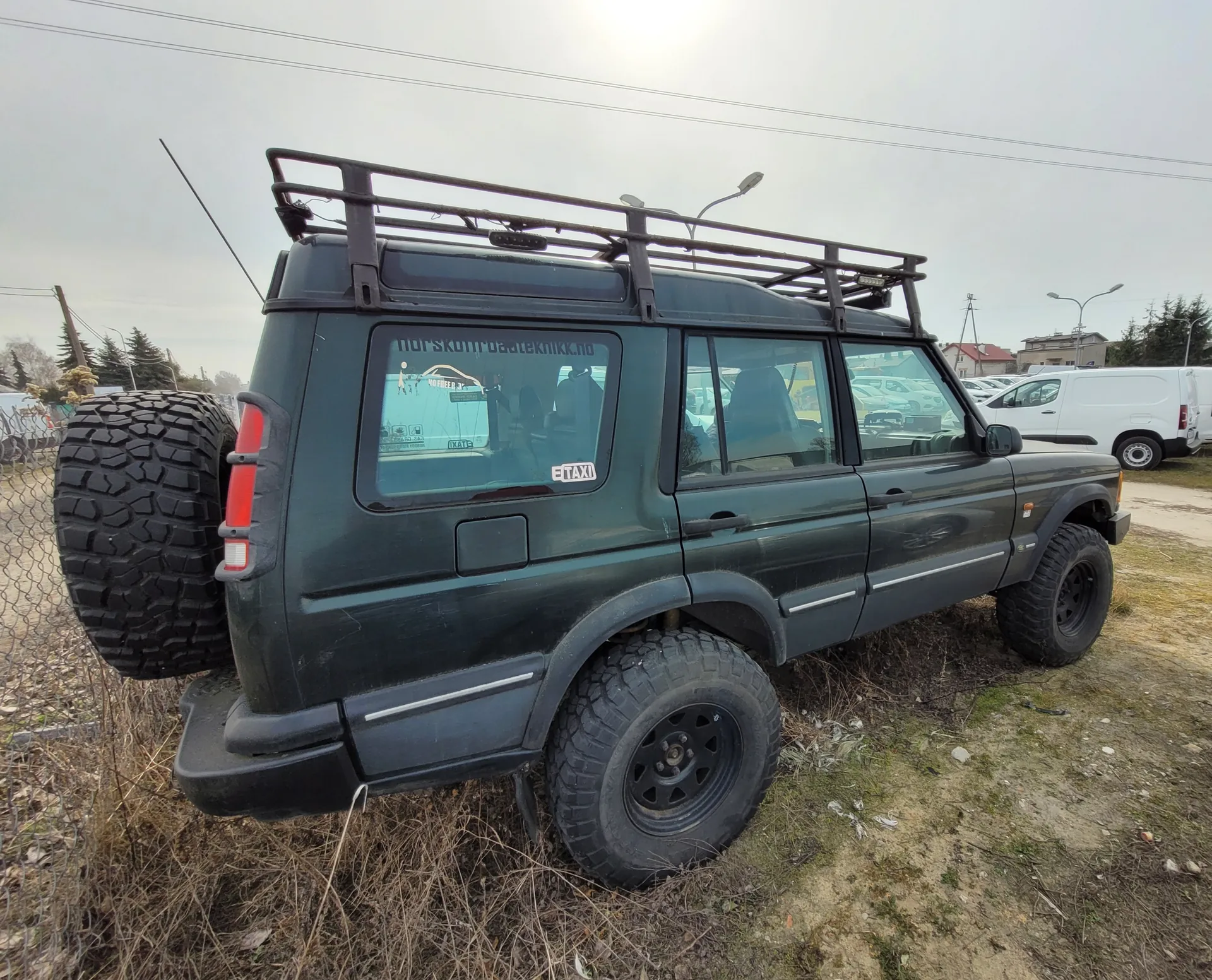Ciemnozielony SUV typu Land Rover Defender z dachowym bagażnikiem i zapasowym kołem na tylnej klapie. Na tylnej szybie widoczny napis 'norskombilteknikk.no' oraz naklejka 'E-TAXI'. Pojazd stoi na trawiastym i częściowo suchym terenie, w tle białe furgony, słupy energetyczne, drzewa i ogrodzenie siatkowe. Niebo jest pochmurne.