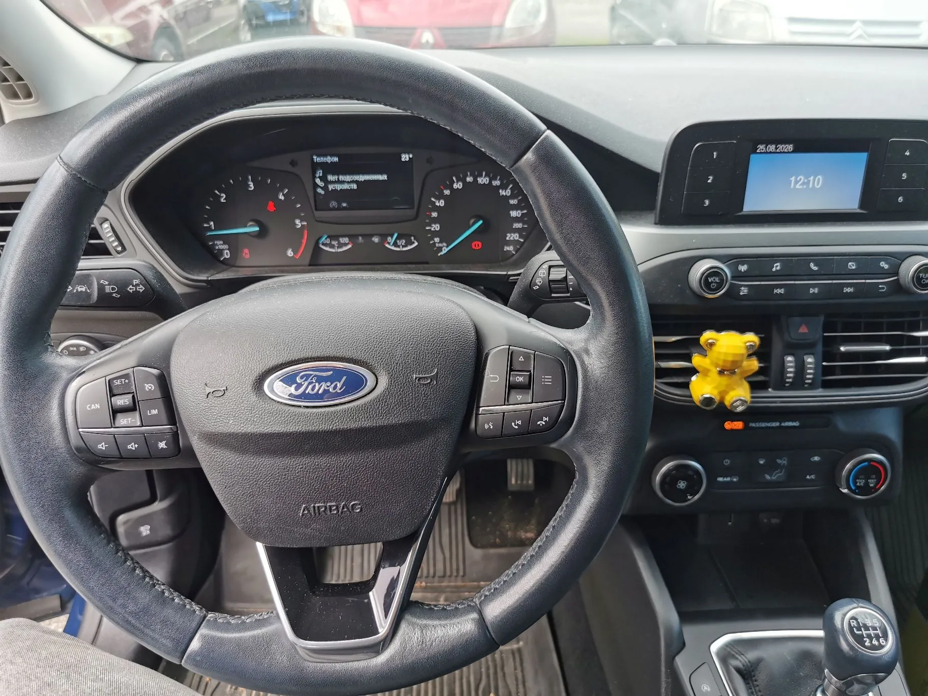 Wnętrze samochodu marki Ford widziane z perspektywy kierowcy. Na pierwszym planie znajduje się czarna, skórzana kierownica z logo Ford i przyciskami sterowania. Za kierownicą widoczny jest panel instrumentów z prędkościomierzem, obrotomierzem i wyświetlaczem informacyjnym w języku rosyjskim. Po prawej stronie znajduje się konsola środkowa z ekranem multimedialnym wyświetlającym godzinę 12:10 i datę 25.08.2026, przyciskami radia oraz nawiewami, do których przyczepiona jest mała, żółta figurka misia. Na dole widoczna jest wajcha zmiany biegów z oznaczeniami przełożeń.