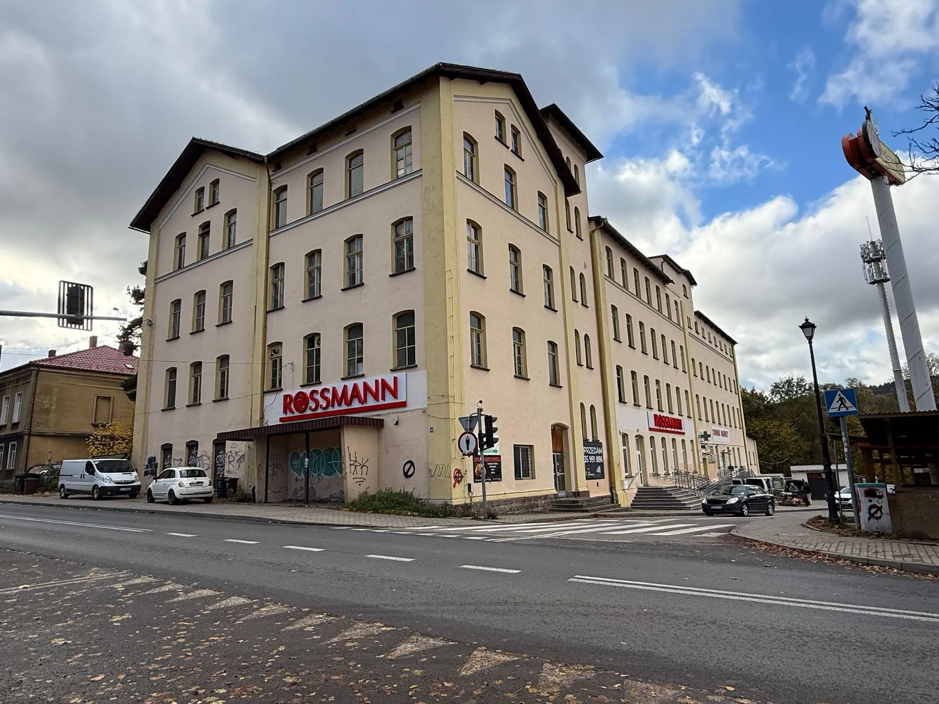 Wielopiętrowy, jasnobeżowy budynek z wieloma oknami, w którym znajduje się drogeria Rossmann. Na fasadzie widnieje czerwony napis 'ROSSMANN'. Budynek stoi przy drodze z pasami dla pieszych. W tle widać zachmurzone niebo z prześwitami błękitu oraz fragmenty zieleni. Po lewej stronie zaparkowane są dwa białe samochody, a po prawej stronie widoczny jest wysoki maszt z reklamą oraz znak drogowy ostrzegający przed przejściem dla pieszych.