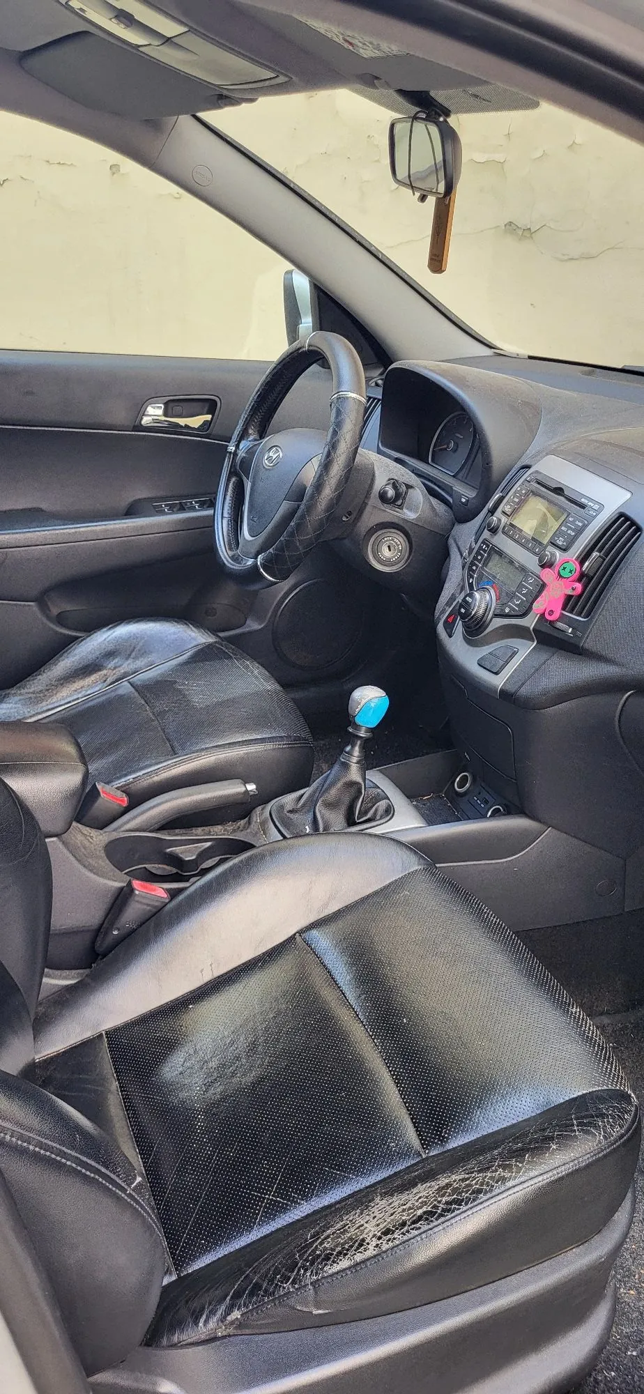 Wnętrze samochodu z perspektywy tylnego siedzenia. Widoczna jest przednia konsola, czarna kierownica z logo Toyoty, deska rozdzielcza z radiem i przyciskami sterowania oraz dwa przednie fotele z czarnej, lekko zużytej skóry. Pomiędzy fotelami znajduje się lewarek zmiany biegów z niebieską gałką. Do konsoli środkowej przyczepiona jest różowa ozdoba w kształcie gwiazdki.