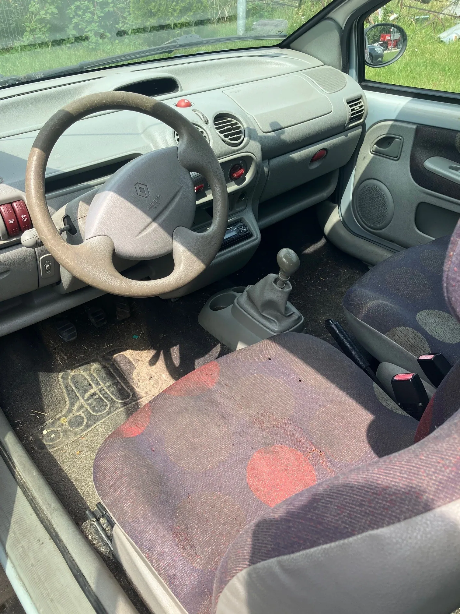 Wnętrze samochodu widziane z perspektywy otwartych drzwi pasażera. Widoczna jest szara deska rozdzielcza, kierownica z logo Renault, lewarek zmiany biegów oraz dwa fotele tapicerowane materiałem w kolorze szarym z czerwonymi kropkami. Na podłodze znajduje się gumowa mata z wypustkami.