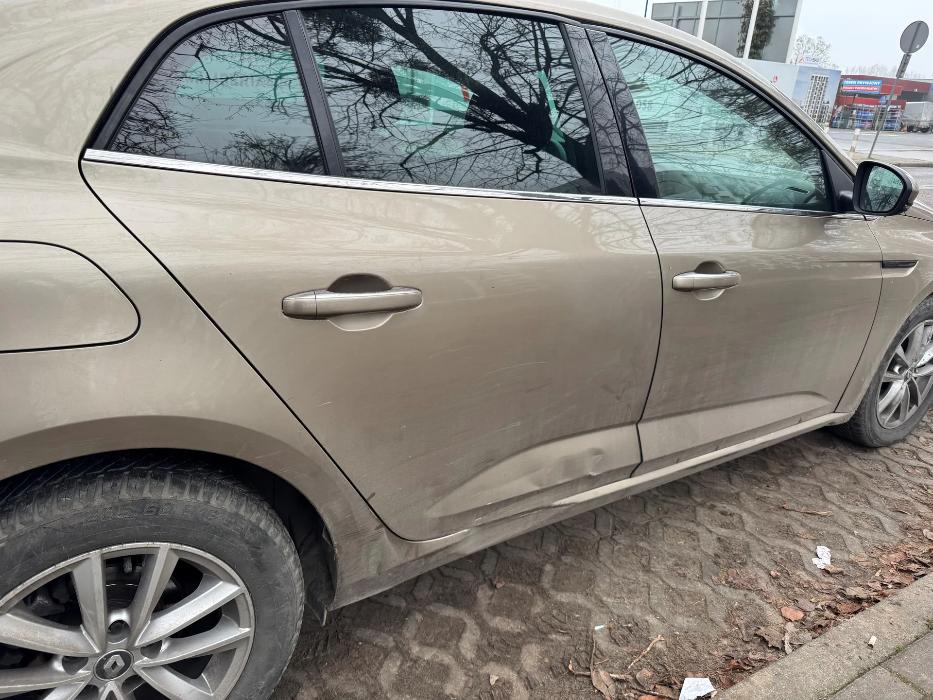 Beżowy samochód osobowy z widocznymi zadrapaniami na karoserii, zaparkowany na brukowanej nawierzchni. Widoczne są drzwi, klamki, felgi oraz odbicia drzew w szybach. W tle budynki i znak drogowy.