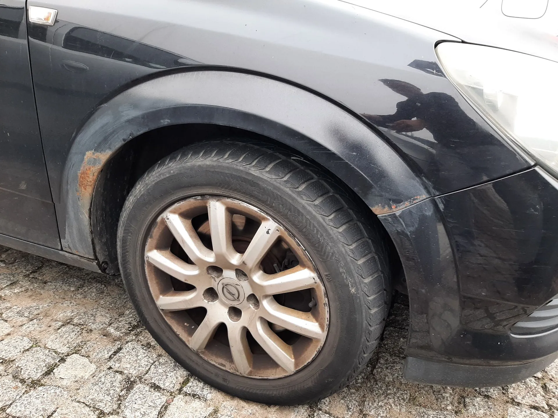 Zbliżenie na przednie prawe koło czarnego samochodu. Felga aluminiowa z logo marki Opel jest zabrudzona pyłem hamulcowym. Na nadkolu, nad oponą, widoczne są ogniska korozji i odpryski lakieru. Samochód stoi na kostce brukowej.