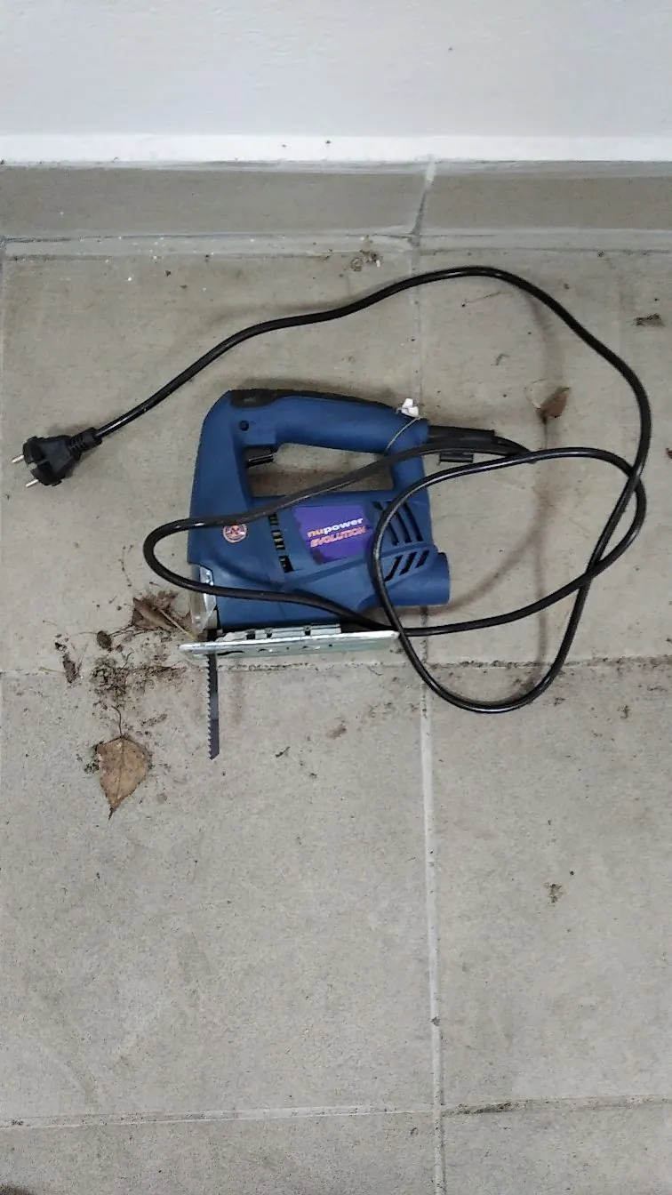 Niebieska wyrzynarka elektryczna marki Nupower Evolution leżąca na szarych płytkach ceramicznych. Urządzenie ma czarny kabel z wtyczką, który jest rozwinięty wokół niego. Obok narzędzia znajdują się kilka suchych liści.