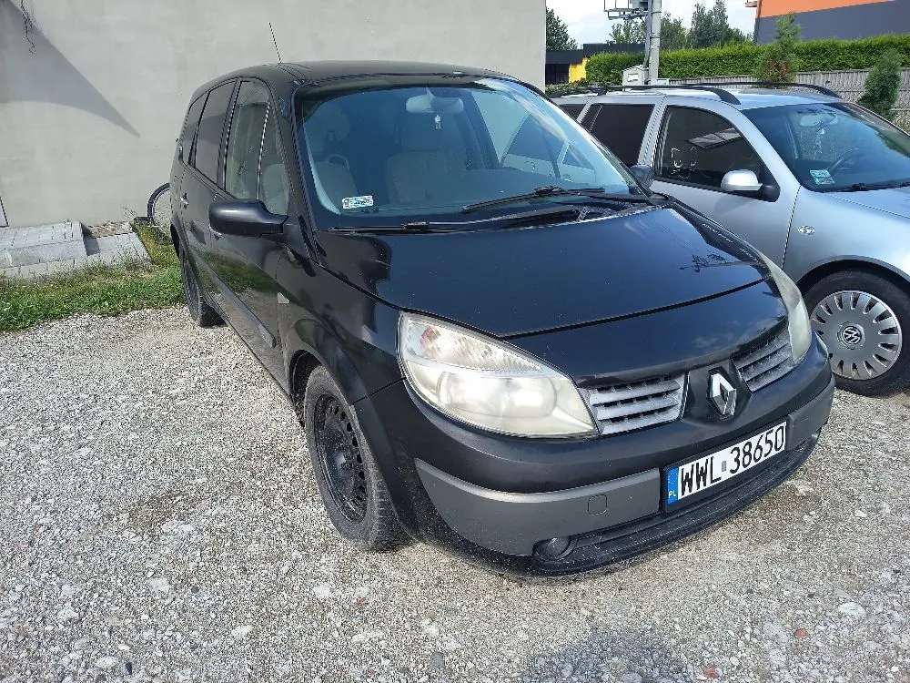 Czarny samochód marki Renault zaparkowany na żwirowym podłożu obok srebrnego samochodu. Widoczna jest przednia część pojazdu z tablicą rejestracyjną o numerze WWL 38650. W tle znajduje się jasna ściana budynku oraz zieleń.