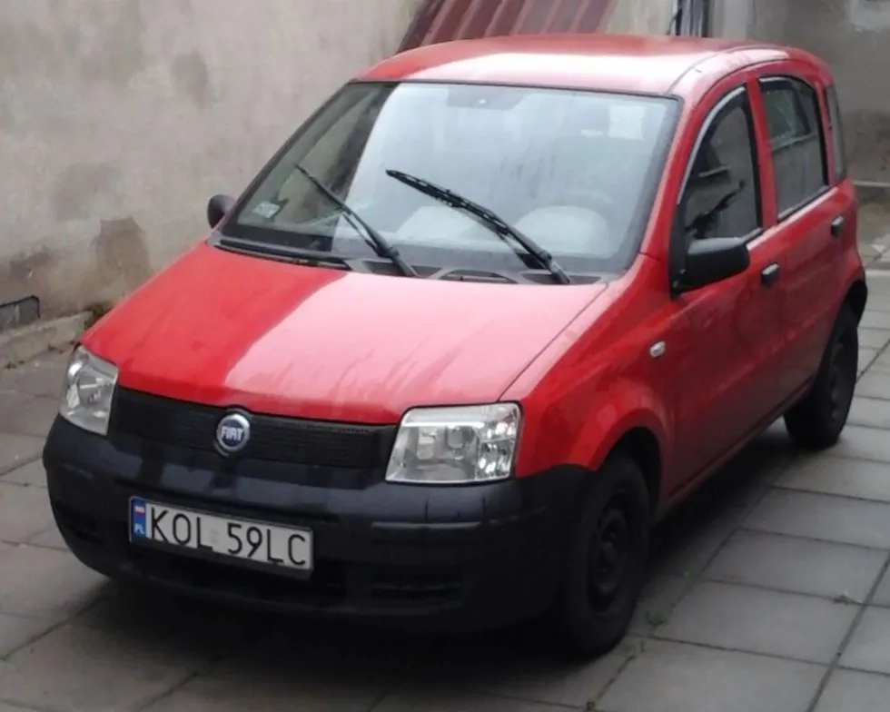 Czerwony samochód marki Fiat zaparkowany na chodniku z szarych płyt betonowych. Pojazd ma czarne zderzaki i czarne felgi. Na przodzie widoczna jest tablica rejestracyjna o treści KOL 59LC.