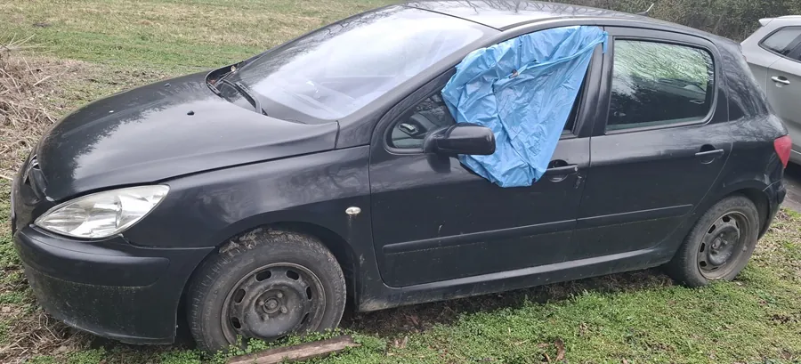 Czarny samochód osobowy zaparkowany na trawie, z nieobecnym przednim lewym kołem, które zastąpiono drewnianą belką. Na oknie pasażera znajduje się niebieska folia. Samochód jest zabrudzony, zwłaszcza koła. W tle widoczna trawa i częściowo inny pojazd. Brak czytelnego tekstu na obrazie.