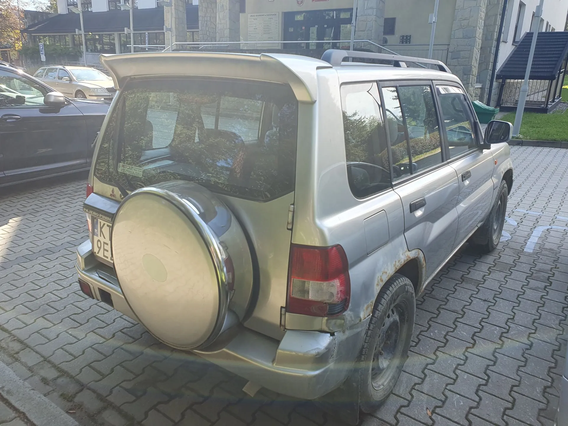 Srebrny samochód terenowy marki Mitsubishi Pajero iO zaparkowany na kostce brukowej. Widok z tyłu i z boku. Pojazd posiada zapasowe koło w twardej osłonie zamontowane na tylnych drzwiach, spojler nad tylną szybą oraz relingi dachowe. Na prawym tylnym błotniku widoczne są ślady korozji. Tablica rejestracyjna zaczyna się od liter KT.