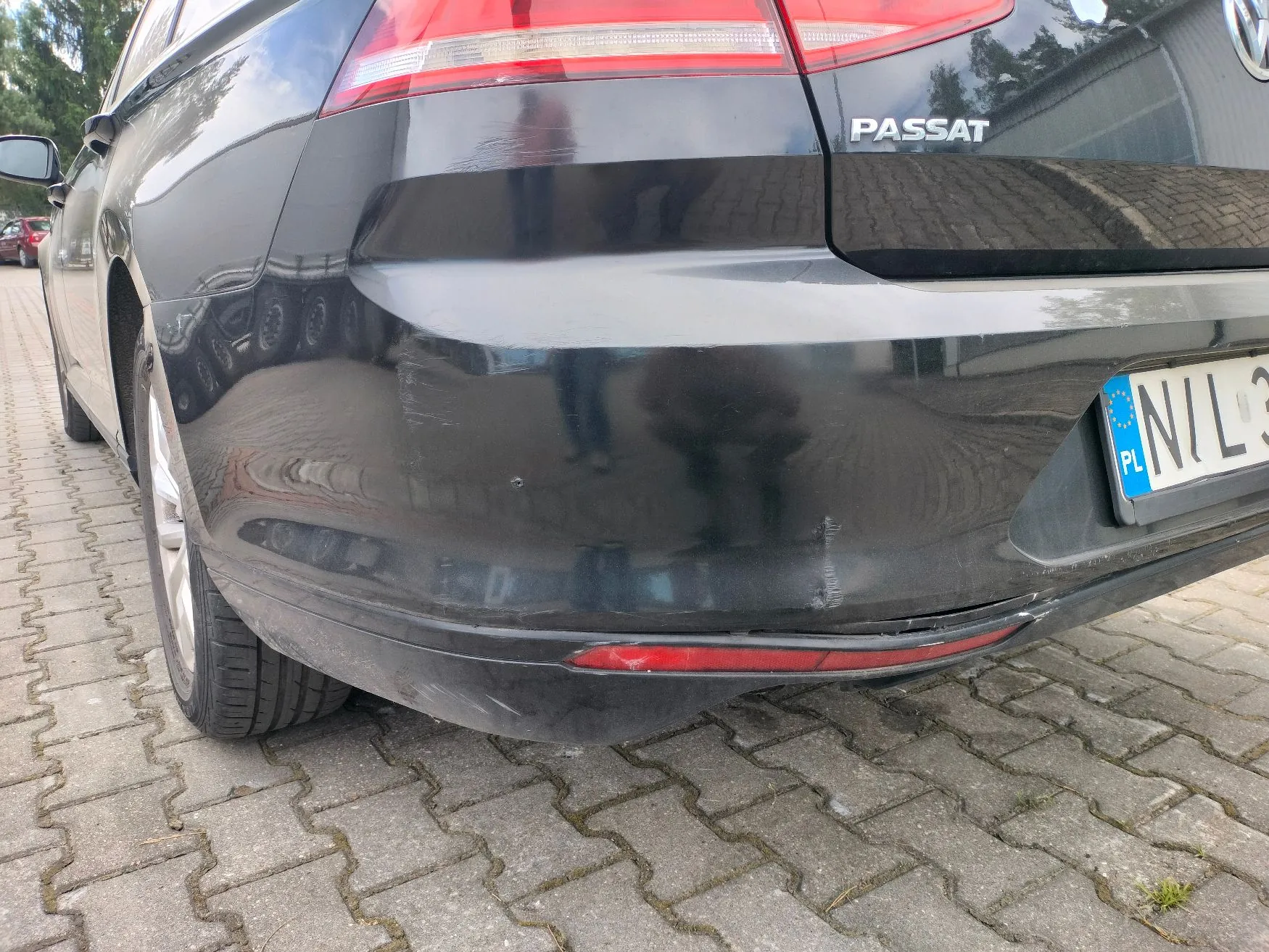 Zdjęcie tylnej części czarnego samochodu marki Volkswagen Passat, zaparkowanego na kostce brukowej. Na tylnym zderzaku widoczne są uszkodzenia w postaci wgnieceń oraz zarysowań lakieru. Widoczny jest fragment tablicy rejestracyjnej z oznaczeniem PL oraz fragment napisu PASSAT na klapie bagażnika.