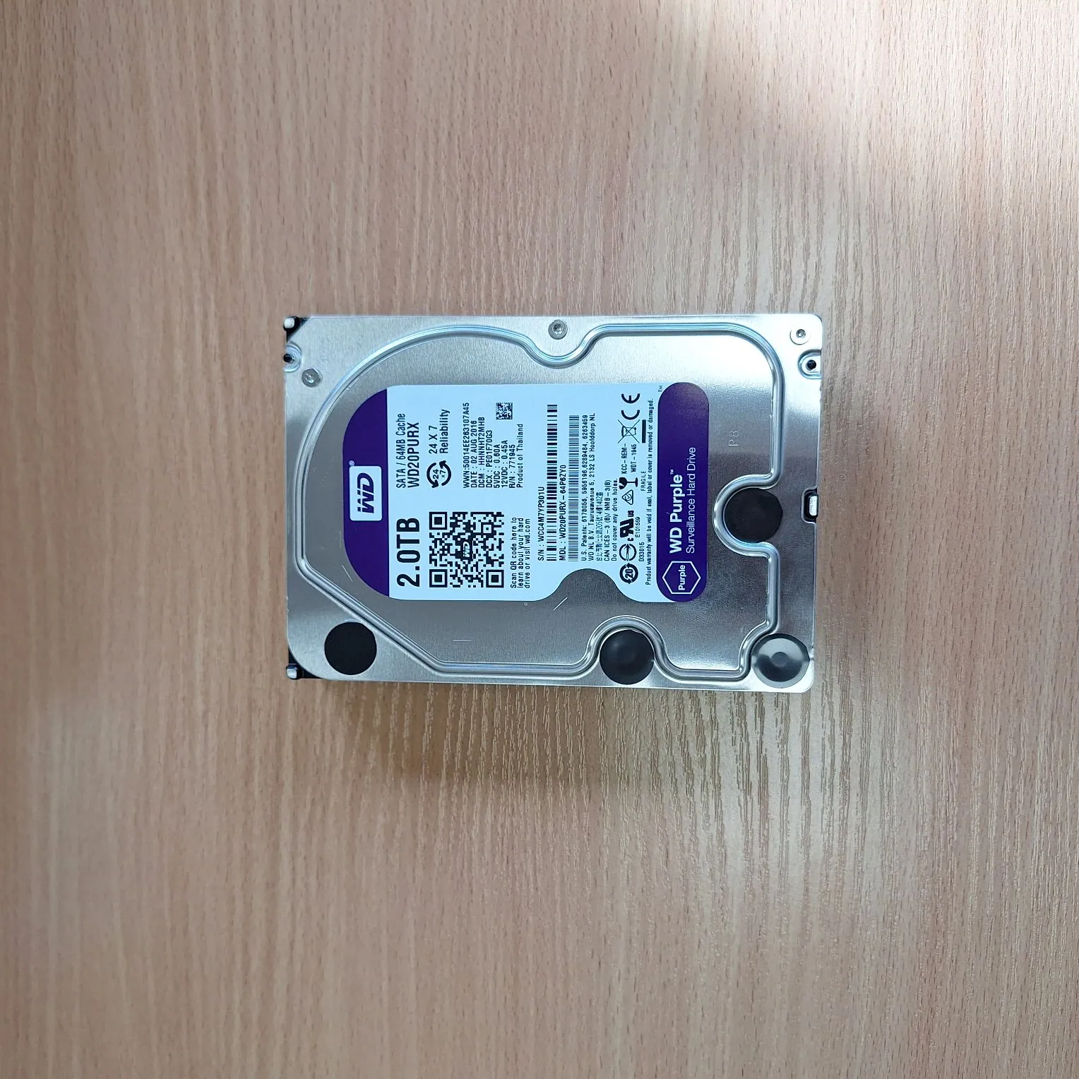 Dysk twardy marki Western Digital (WD) o pojemności 2.0TB, model WD20PURX z serii WD Purple Surveillance Hard Drive. Urządzenie leży na jasnym, drewnianym blacie. Na srebrnej obudowie znajduje się biała etykieta z logo WD, kodem QR, kodami kreskowymi oraz specyfikacją techniczną i certyfikatami.