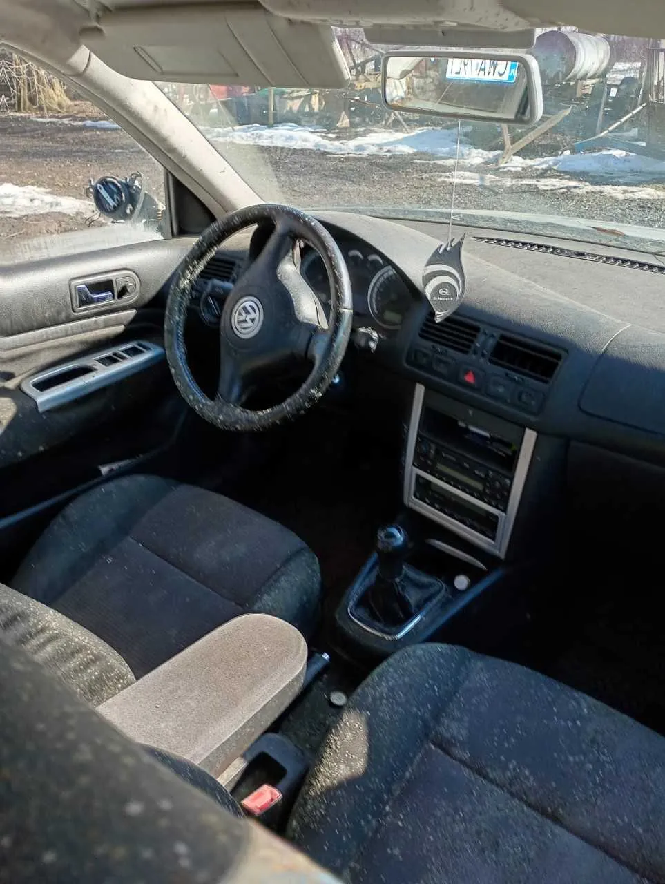 Wnętrze samochodu marki Volkswagen z widocznym kierownicą z logo VW, czarnymi siedzeniami pokrytymi kurzem, centralną konsolą z manualną skrzynią biegów i odtwarzaczem, lusterkiem wstecznym z tablicą rejestracyjną LKV 4WJ. Przez przednią szybę widoczny zewnętrzny krajobraz z resztkami śniegu i beczką na tle.