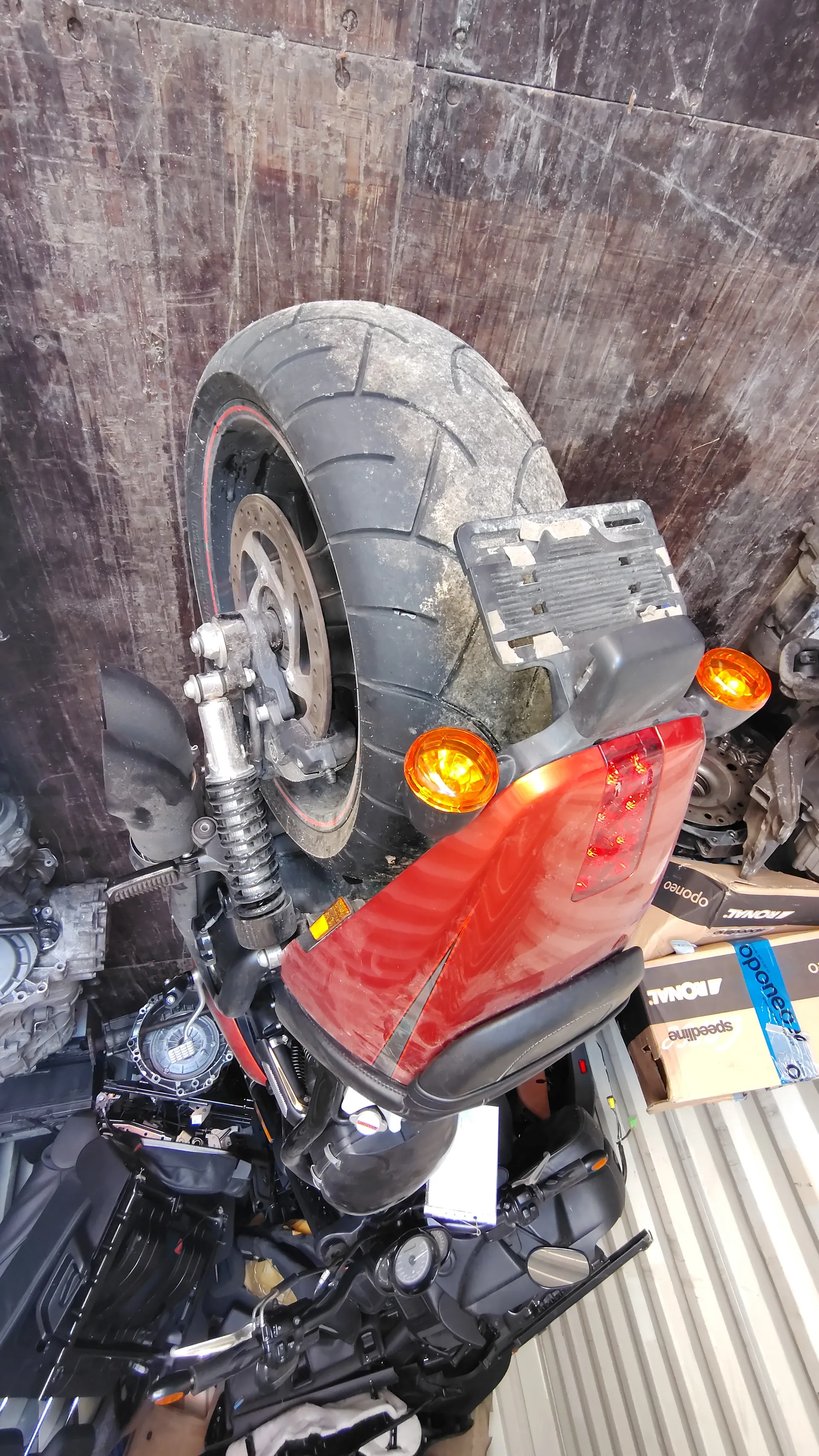 Czerwony motocykl widziany od tyłu z dużym, brudnym kołem tylnym, pomarańczowymi kierunkowskazami i czerwonym światłem tylnym. Puste miejsce na tablicę rejestracyjną. W tle pudełka z widocznym tekstem 'oponeo' i 'speedline', części mechaniczne oraz podłoga z drewnianych desek i metalowa ściana.
