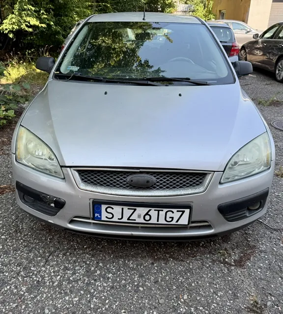 Srebrny samochód osobowy marki Ford widziany od przodu. Pojazd posiada polską tablicę rejestracyjną o numerze SJZ 6TG7. Auto stoi na asfaltowym podłożu, w tle widoczne są inne zaparkowane samochody oraz zielone drzewa.