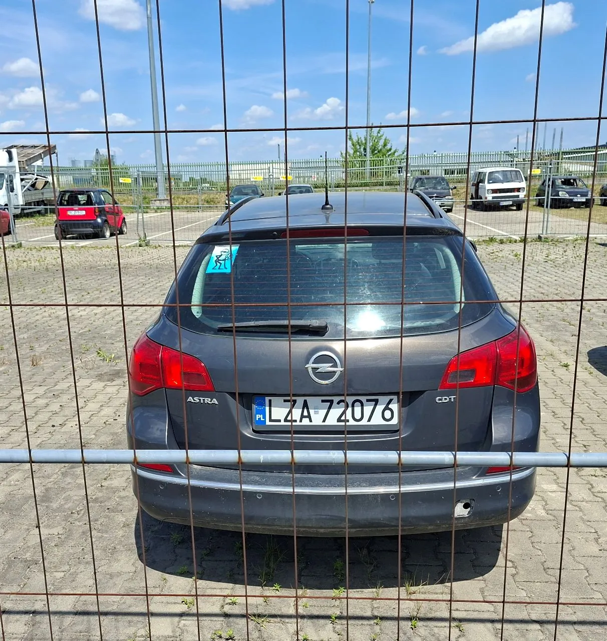 Widok z tyłu szarego samochodu marki Opel Astra CDT, zaparkowanego na placu z kostki brukowej. Samochód ma polską tablicę rejestracyjną o numerze LZA 72076. Zdjęcie zostało wykonane przez metalowy płot z siatki, który znajduje się na pierwszym planie. W tle widoczne są inne zaparkowane samochody, w tym czerwony samochód po lewej stronie, oraz niebieskie niebo z białymi chmurami.