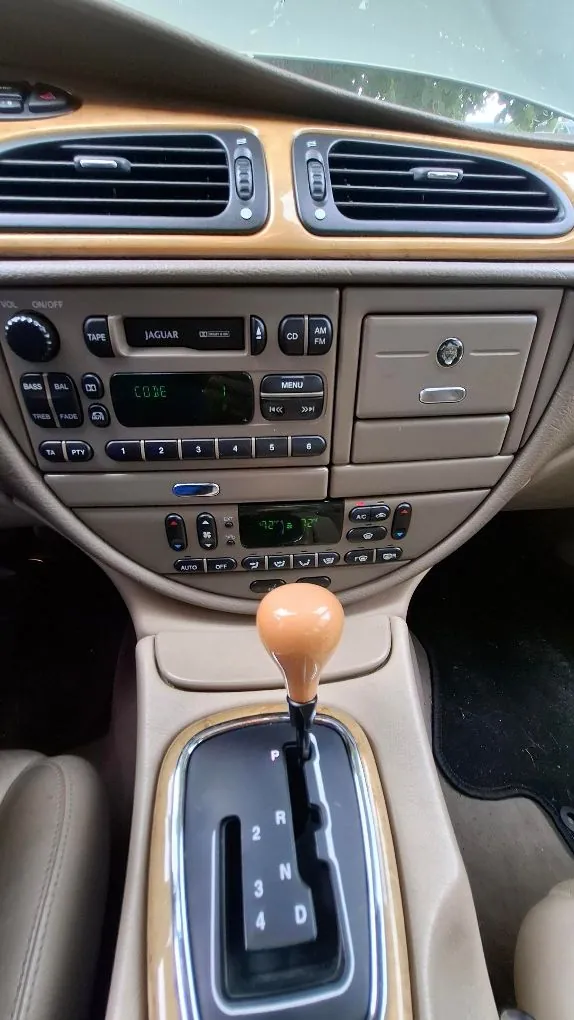 Wnętrze samochodu marki Jaguar, widok na konsolę środkową. W górnej części znajdują się dwa poziome nawiewy. Poniżej znajduje się radio z napisem 'JAGUAR', wyświetlaczem z zielonym tekstem 'CODE' oraz przyciskami sterującymi. Pod radiem znajduje się panel klimatyzacji z cyfrowym wyświetlaczem temperatury. Na dole widoczna jest wajcha zmiany biegów automatycznej skrzyni z oznaczeniami P, R, N, D oraz cyframi 2, 3, 4, osadzona w obudowie z elementami drewnianymi.