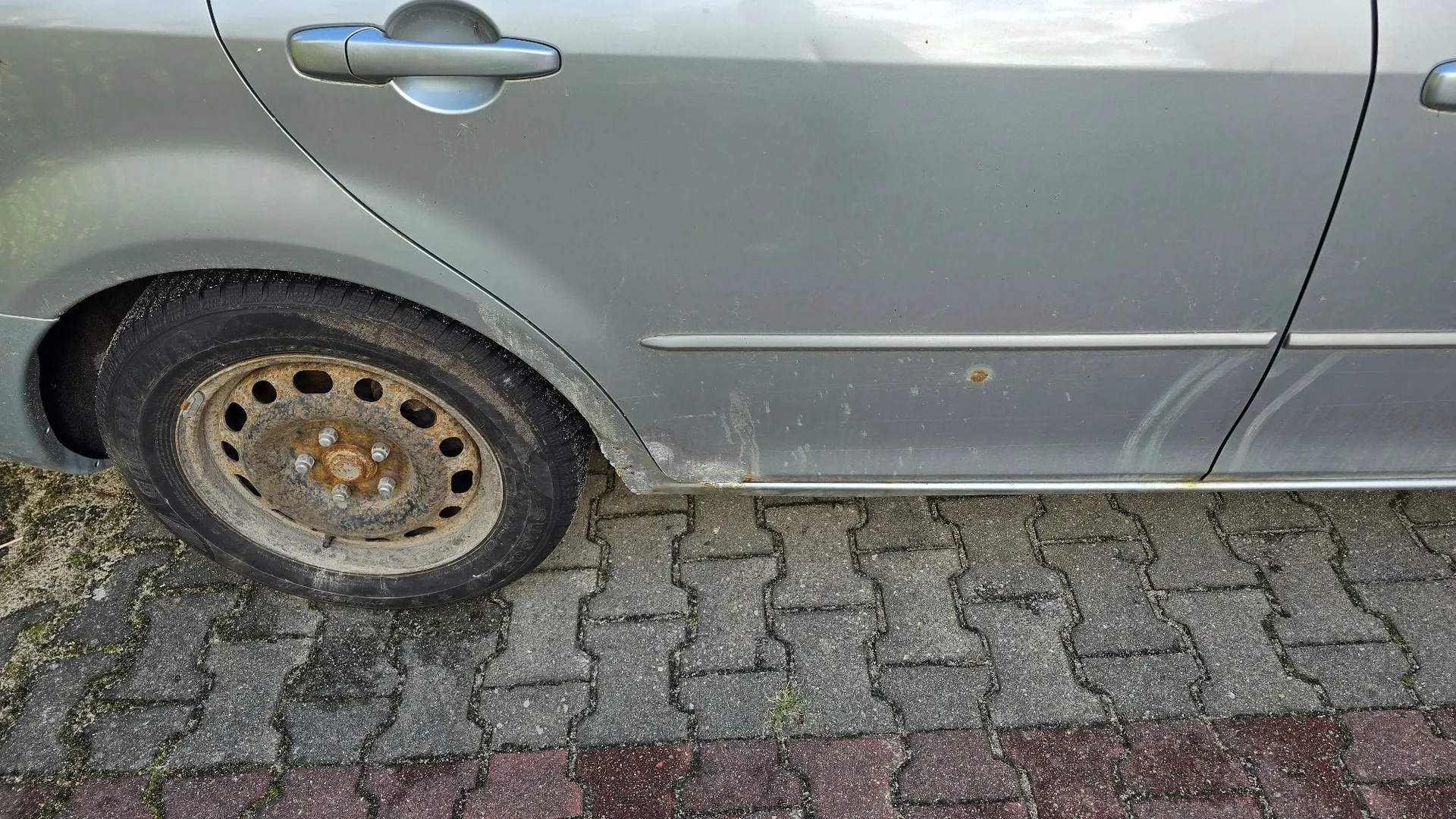 Zbliżenie na dolną część srebrnego samochodu, pokazujące tylne koło z brudną, stalową felgą oraz fragment drzwi i nadkola. Na dolnej krawędzi nadwozia widoczne są ślady korozji i odpryski lakieru. Samochód stoi na szarej kostce brukowej z czerwonym pasem na dole.