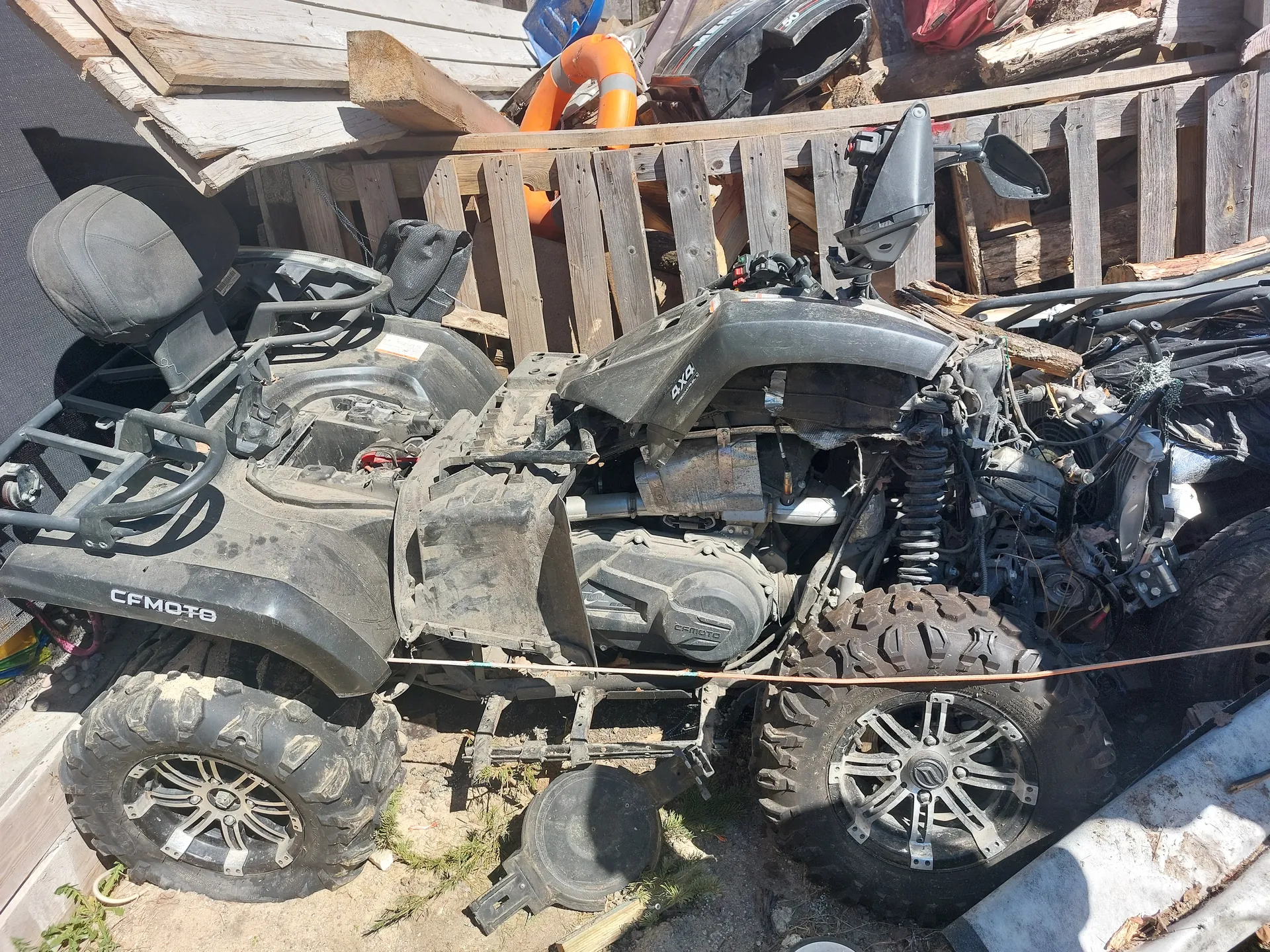 Zniszczony czarny quad marki CFMOTO z odsłoniętym silnikiem i brakującą przednią częścią. Widoczne duże, błotne opony z metalowymi felgami. W tle drewniane deski, pomarańczowy pierścień ratunkowy, niebieski pojemnik, czerwona tkanina oraz różne przedmioty. Na boku quadu widnieje napis CFMOTO. Podłoże ziemne z fragmentami trawy.