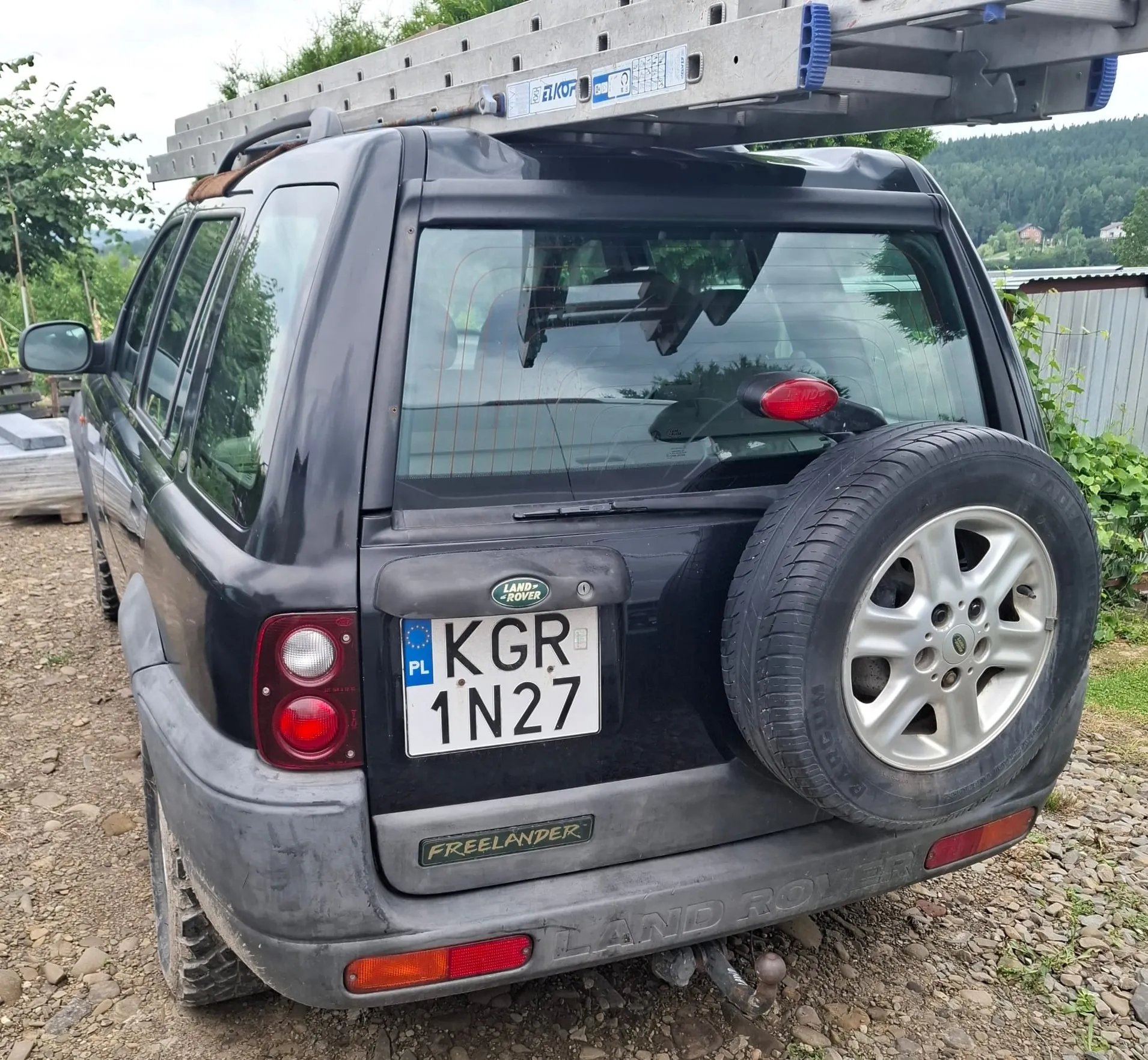 Czarny samochód terenowy Land Rover Freelander zaparkowany na żwirowym podłożu. Na dachu pojazdu zamontowana jest długa, aluminiowa drabina marki ELKOP. Na tylnej klapie znajduje się koło zapasowe, logo Land Rover oraz polska tablica rejestracyjna o numerze KGR 1N27. Na dolnym zderzaku widnieje napis FREELANDER oraz LAND ROVER.
