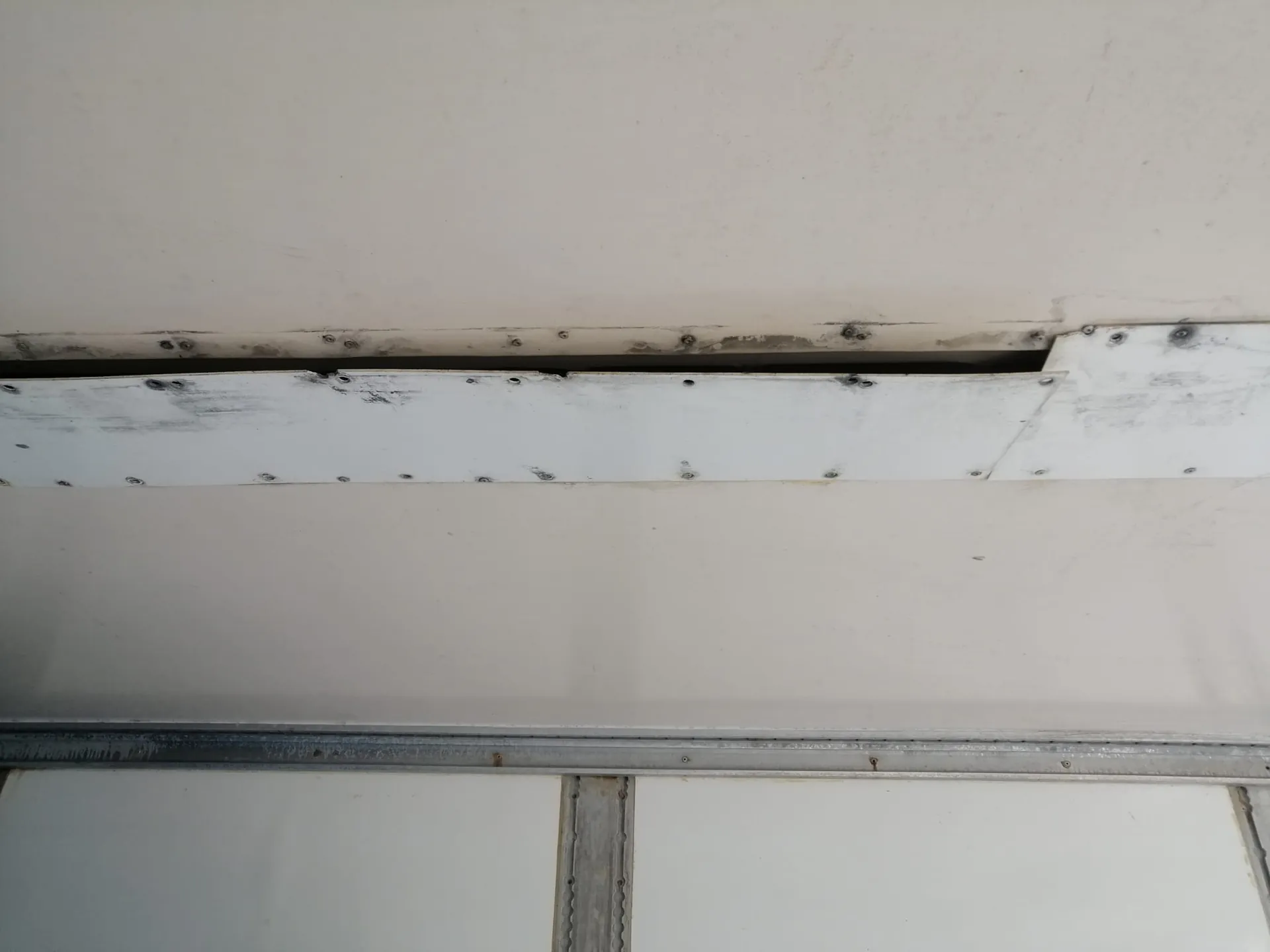 Zbliżenie na białą, metalową konstrukcję z widocznymi nitami i szczeliną wzdłuż krawędzi paneli. Powierzchnia jest lekko zabrudzona, z widocznymi śladami zużycia i ciemniejszymi punktami w miejscach łączeń.