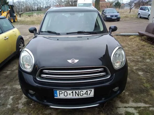 Czarny samochód Mini Cooper z przodu, z widoczną tablicą rejestracyjną PL PO 1NG47. Po lewej stronie żółty samochód, w tle inne pojazdy (ciemny samochód, srebrny van) oraz żółty pojazd budowlany. Tło: trawiasto-ziemista powierzchnia, budynki i drzewa.