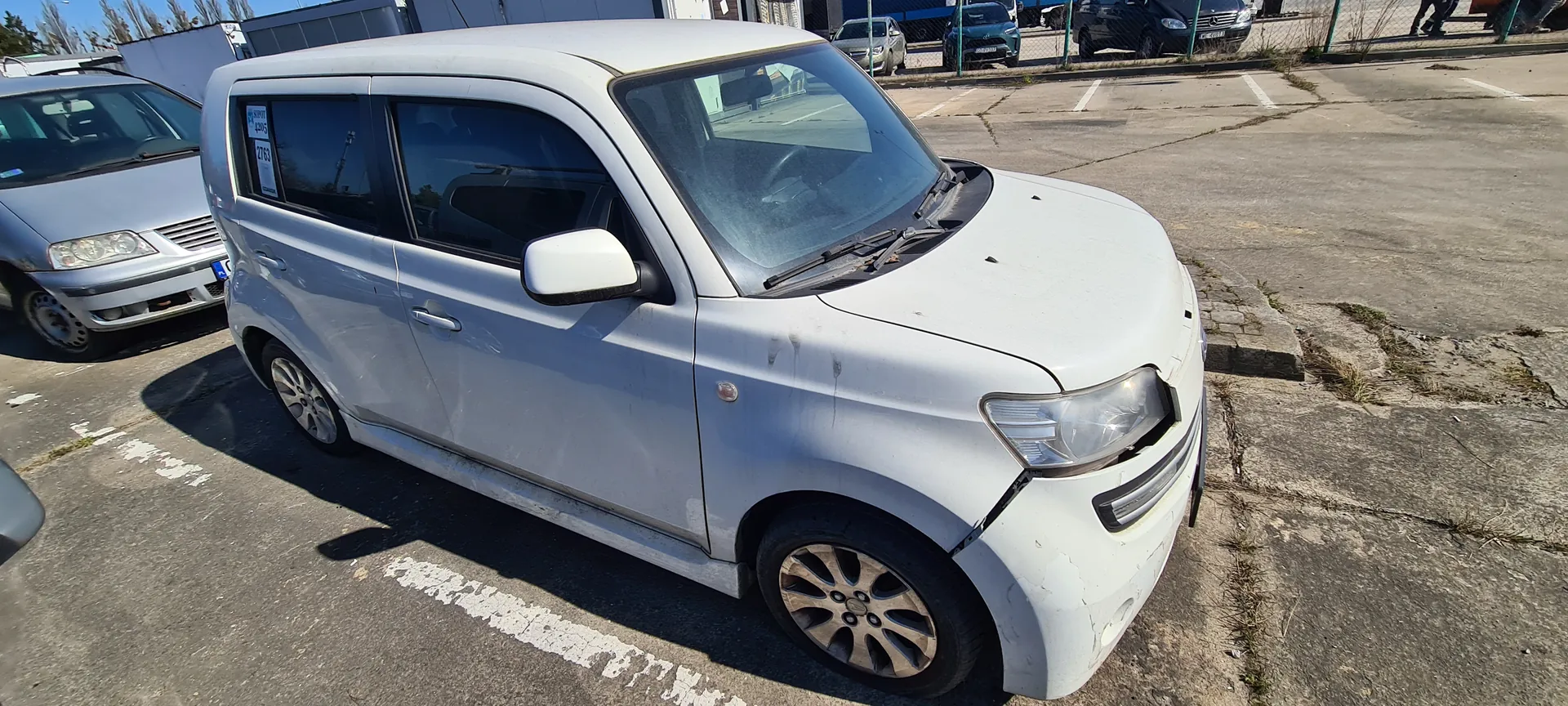 Biały samochód osobowy zaparkowany na betonowym placu parkingowym. Pojazd ma widoczne uszkodzenia na przednim prawym zderzaku i karoserii. Na tylnym oknie widoczny naklejony znak z cyframi 2763. W tle inne zaparkowane samochody, ogrodzenie i budynki. Słoneczny dzień, cienie wskazują na jasne oświetlenie. Linie parkingowe na asfalcie są białe, a powierzchnia placu ma pęknięcia i miejscami rośnie trawa.