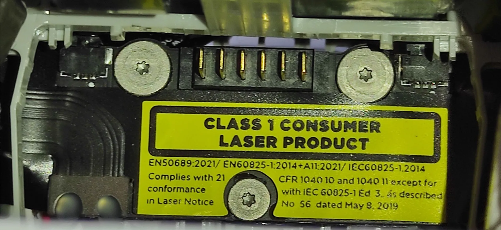 Zbliżenie na żółtą etykietę ostrzegawczą umieszczoną na czarnym elemencie urządzenia elektronicznego. Na etykiecie widnieje napis: 'CLASS 1 CONSUMER LASER PRODUCT'. Poniżej znajduje się mniejszy tekst: 'EN50689:2021 / EN60825-1:2014+A11:2021 / IEC60825-1:2014 Complies with 21 conformance in Laser Notice CFR 1040 10 and 1040 11 except for with IEC 60825-1 Ed 3 as described No 56 dated May 8, 2019'. Nad etykietą widoczne są dwa srebrne wkręty z łbami w kształcie gwiazdy oraz złote piny złącza elektrycznego.