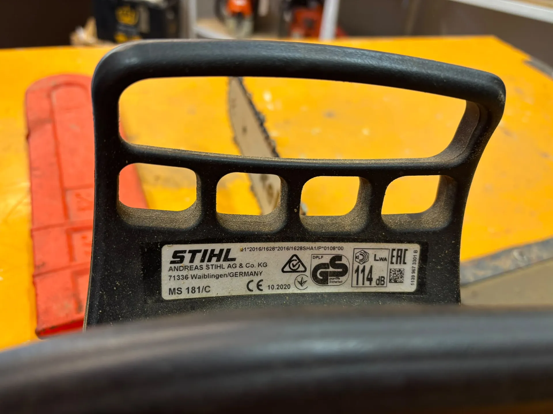 Zbliżenie na czarny uchwyt piły spalinowej marki STIHL. Na uchwycie znajduje się srebrna tabliczka znamionowa z tekstem: 'STIHL ANDREAS STIHL AG & Co. KG 71336 Waiblingen/GERMANY MS 181/C', a także oznaczeniami CE, datą 10.2020, symbolem GS oraz poziomem hałasu '114 dB'. W tle widoczna jest żółta powierzchnia oraz fragment czerwonego elementu urządzenia.