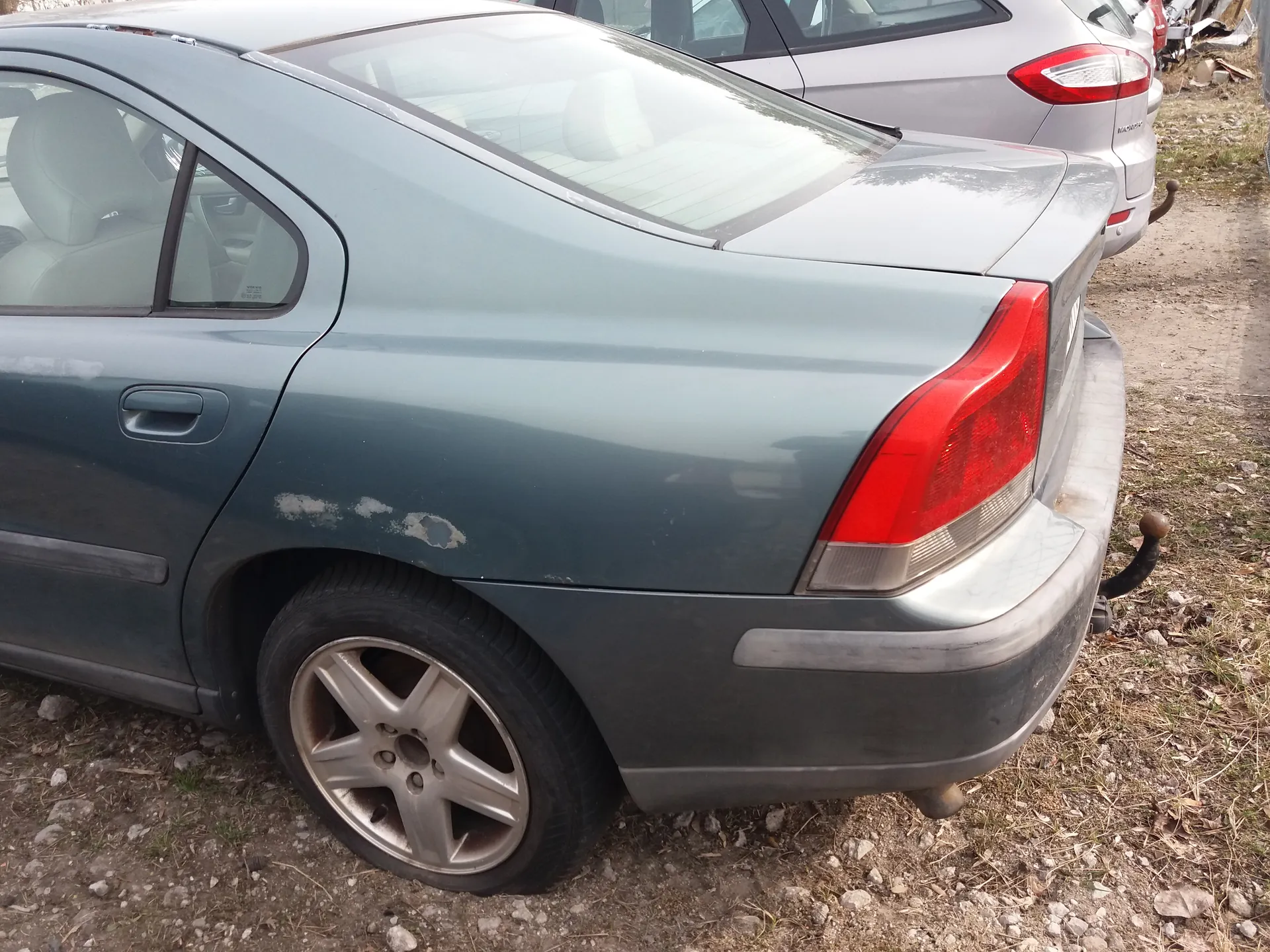 Zdjęcie przedstawia tylną część szarego samochodu osobowego typu sedan. Widoczne są uszkodzenia na boku nadwozia w postaci zadrapań i otworu. Samochód ma czerwone światło tylnego reflektora, srebrne felgi oraz zarysowaną tablicę rejestracyjną. W tle znajduje się inny srebrny samochód osobowy. Podwozie stoi na podłożu z żwirem i suchą trawą.