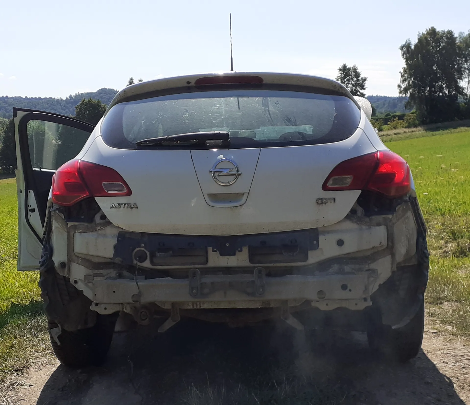 Tylny widok białego samochodu marki Opel Astra, któremu brakuje tylnego zderzaka, odsłaniając metalowe elementy podwozia. Lewe tylne drzwi są otwarte. Samochód stoi na piaszczystej drodze w otoczeniu zielonej trawy i drzew w tle.