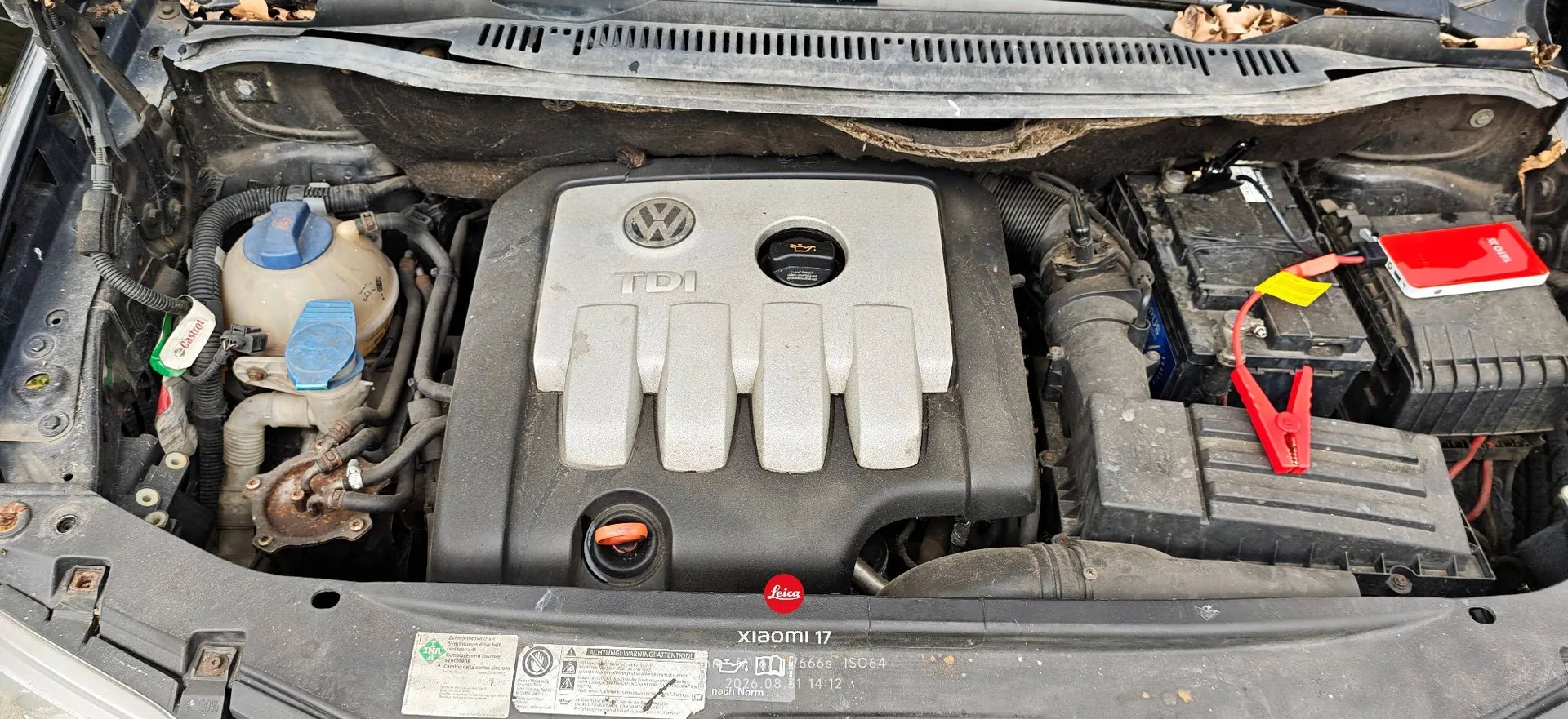 Widok z góry na komorę silnika samochodu marki Volkswagen. W centrum znajduje się plastikowa osłona silnika z logo VW i napisem TDI. Po lewej stronie widoczny jest zbiornik z niebieską zakrętką, a po prawej akumulator z podłączonym czerwonym kablem rozruchowym i białym urządzeniem typu powerbank/booster z czerwonymi elementami. W dolnej części zdjęcia widoczny jest fragment nadwozia z naklejkami technicznymi oraz napisem 'xiaomi 17' i 'leica'.