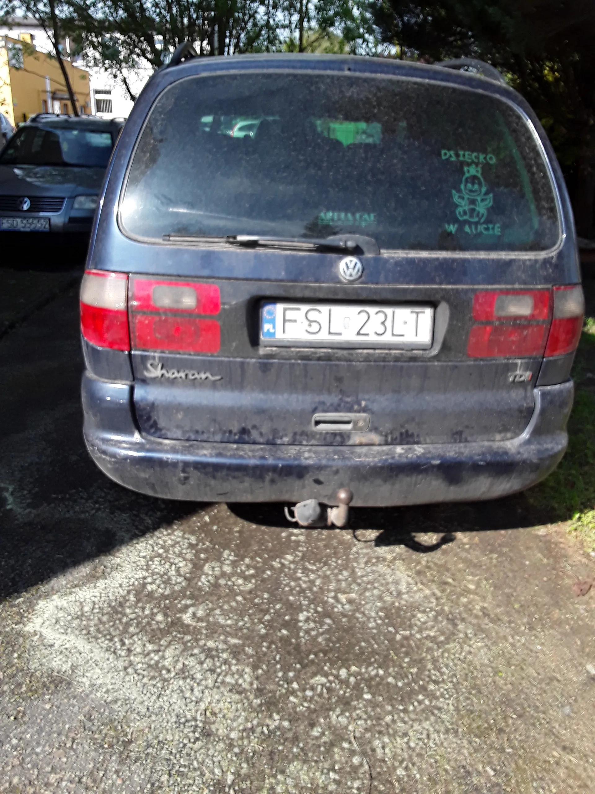 Tył ciemnoszarego samochodu marki Volkswagen model Sharan z tablicą rejestracyjną FSL 23LT. Na szybie tylnej widoczne naklejki, w tym z napisem DZIECKO W 410CE oraz zielonym rysunkiem. Samochód stoi na żwirowej powierzchni, w tle drzewa i fragment innego pojazdu. Czerwone światła tylne oraz logo marki są widoczne na bagażniku.