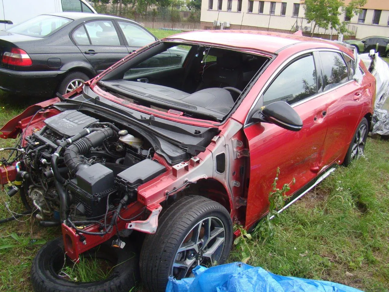 Zdjęcie uszkodzonego, czerwonego samochodu osobowego typu hatchback, który znajduje się na trawiastym terenie. Pojazd jest poważnie rozbity: brakuje przedniego zderzaka, maski oraz przedniej szyby, co odsłania elementy silnika i układu chłodzenia. Prawe drzwi i błotnik są wgniecione. W tle widoczny jest czarny samochód oraz fragmenty budynków.