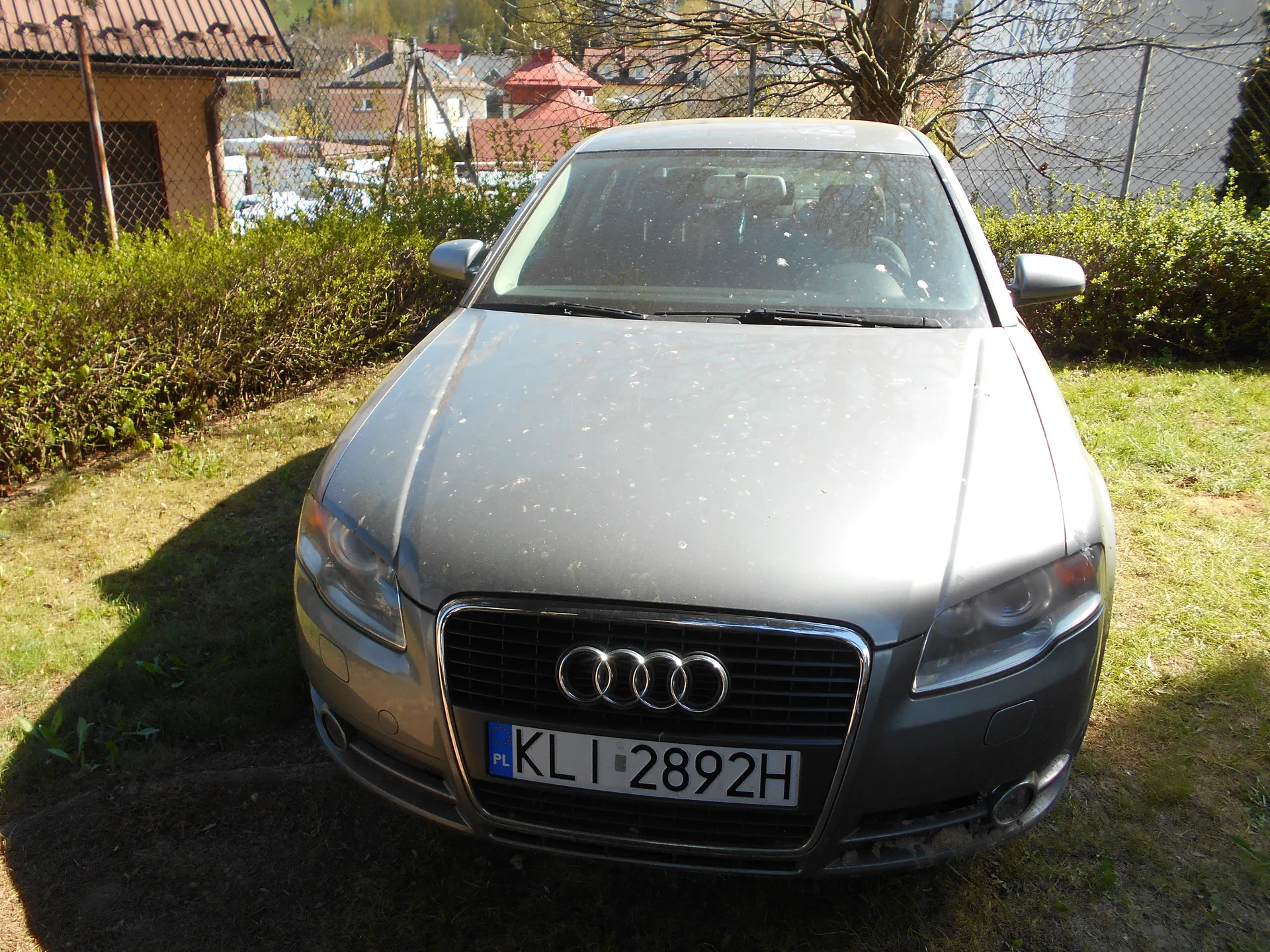 Srebrny samochód marki Audi z przodu, z widoczną tablicą rejestracyjną PL KLI 2892H. Samochód stoi na trawie, w tle siatka ogrodzeniowa, krzewy, domy z czerwonymi dachami oraz drzewo bez liści. Na masce widoczne są ślady brudu. W tle również inne pojazdy i zabudowania.