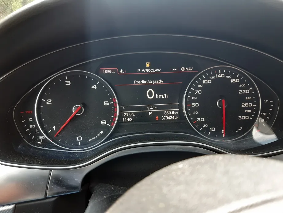 Zbliżenie na deskę rozdzielczą samochodu. Widoczne są dwa analogowe wskaźniki: po lewej obrotomierz, po prawej prędkościomierz z maksymalną wartością 300 km/h. Pomiędzy nimi znajduje się cyfrowy wyświetlacz pokazujący prędkość 0 km/h, napis 'Prędkość jazdy', temperaturę +21.0°C, godzinę 11:53 oraz przebieg 379434 km. Na górnym pasku wyświetlacza widnieje napis 'WROCLAW' oraz ikona stacji paliw i zasięg 50 km.
