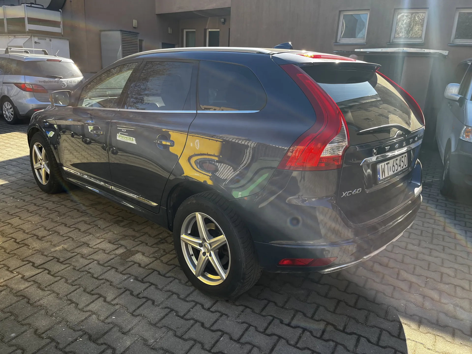 Ciemnoszary samochód Volvo XC60 zaparkowany na brukowanej powierzchni. Widoczny tył i bok pojazdu z tablicą rejestracyjną WT 65450. W tle budynki z oknami oraz inne zaparkowane samochody. Słoneczne oświetlenie tworzy refleksy na karoserii.