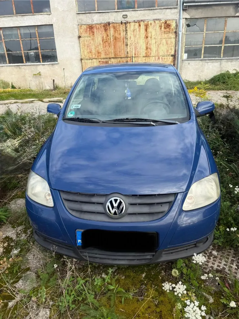 Niebieski samochód marki Volkswagen zaparkowany przodem do widza na trawiastym terenie przed starym, zniszczonym budynkiem przemysłowym z rdzawymi metalowymi drzwiami i wybitymi szybami w oknach.