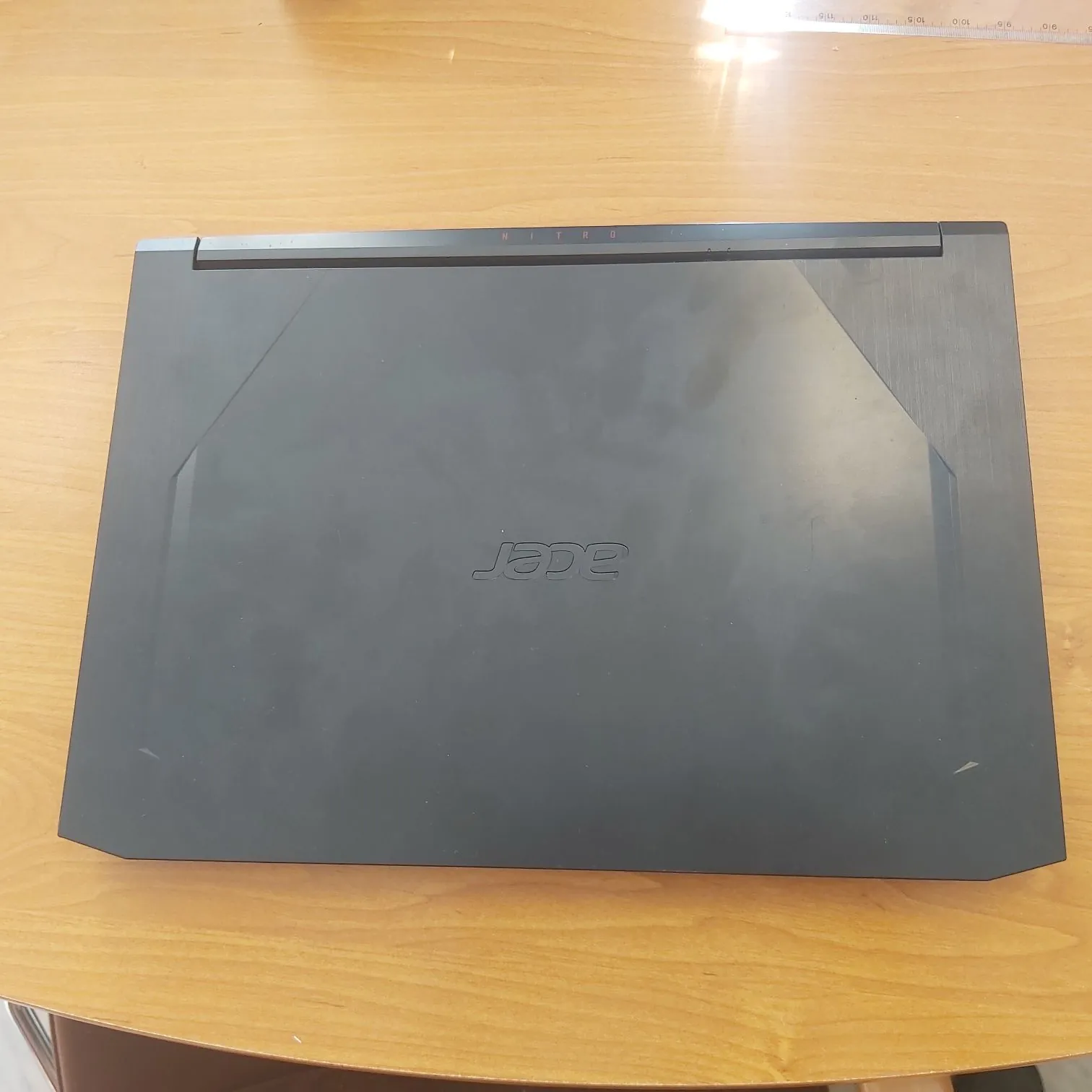 Zamknięty, czarny laptop marki Acer leżący na drewnianym stole. Na pokrywie laptopa znajduje się centralnie umieszczony napis 'acer'. Po prawej krawędzi obudowy widnieje pionowy napis 'NITRO'.