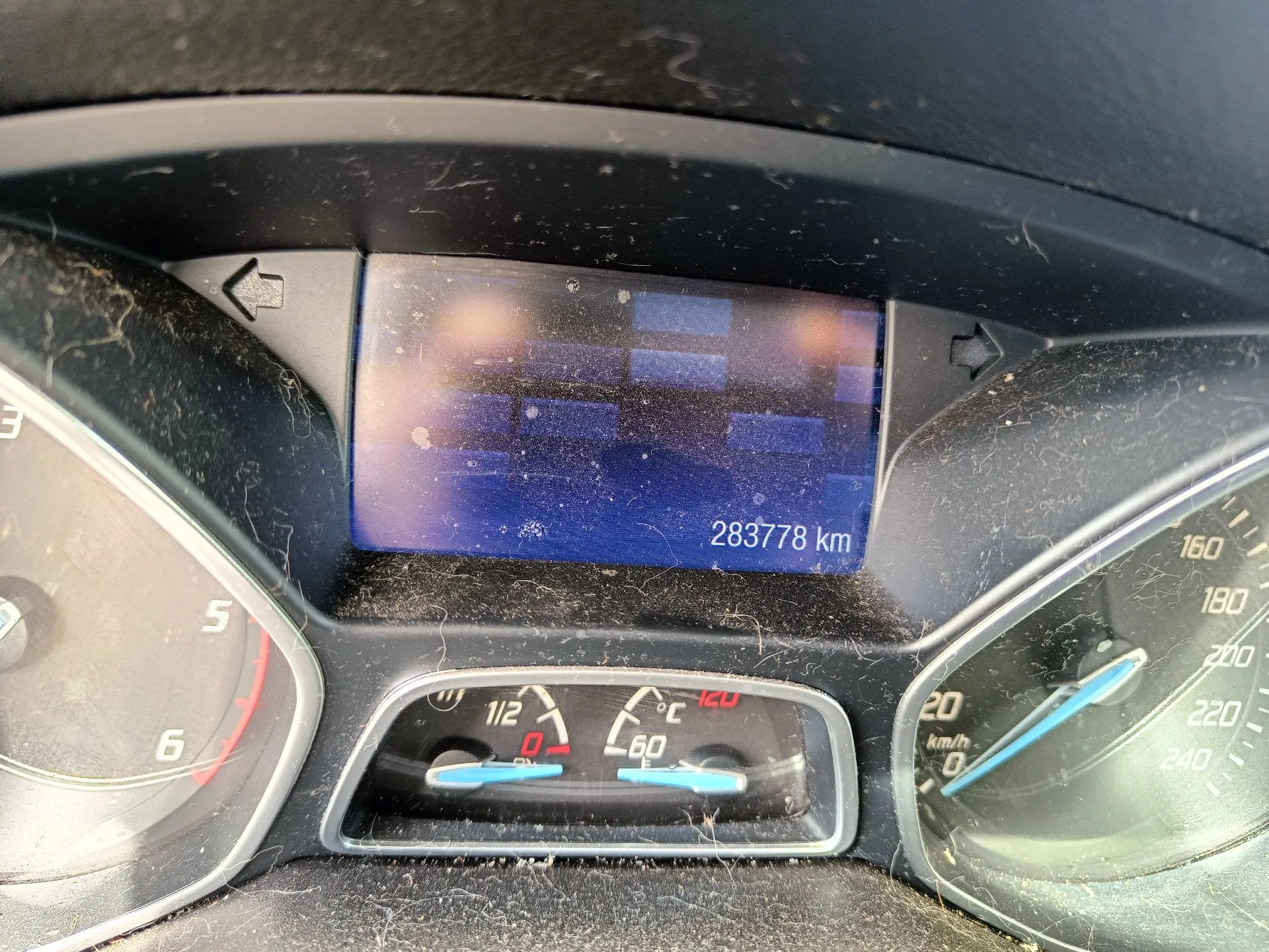 Zbliżenie na deskę rozdzielczą samochodu. W centrum znajduje się cyfrowy wyświetlacz z niebieskim tłem, na którym widnieje przebieg pojazdu: 283778 km. Poniżej ekranu znajdują się analogowe wskaźniki temperatury cieczy chłodzącej oraz poziomu paliwa. Po prawej stronie widoczny jest fragment prędkościomierza z podziałką do 240 km/h. Powierzchnie deski rozdzielczej są zakurzone i pokryte drobnymi włóknami.