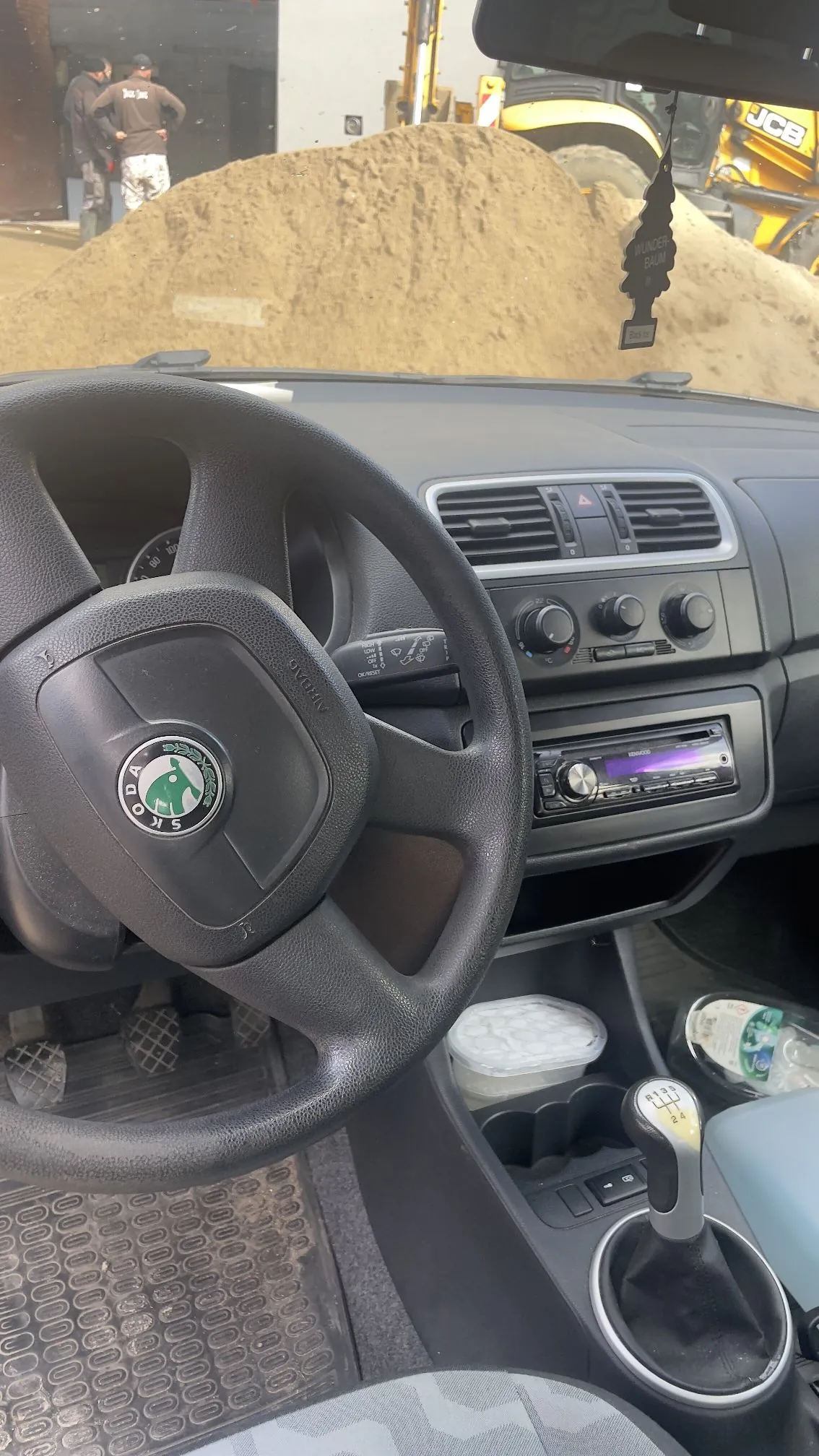 Widok z wnętrza samochodu marki Skoda, pokazujący kierownicę z logo producenta, deskę rozdzielczą z nawiewami i radiem oraz lewarek zmiany biegów. Przez przednią szybę widoczna jest pryzma piasku oraz żółta koparka JCB.